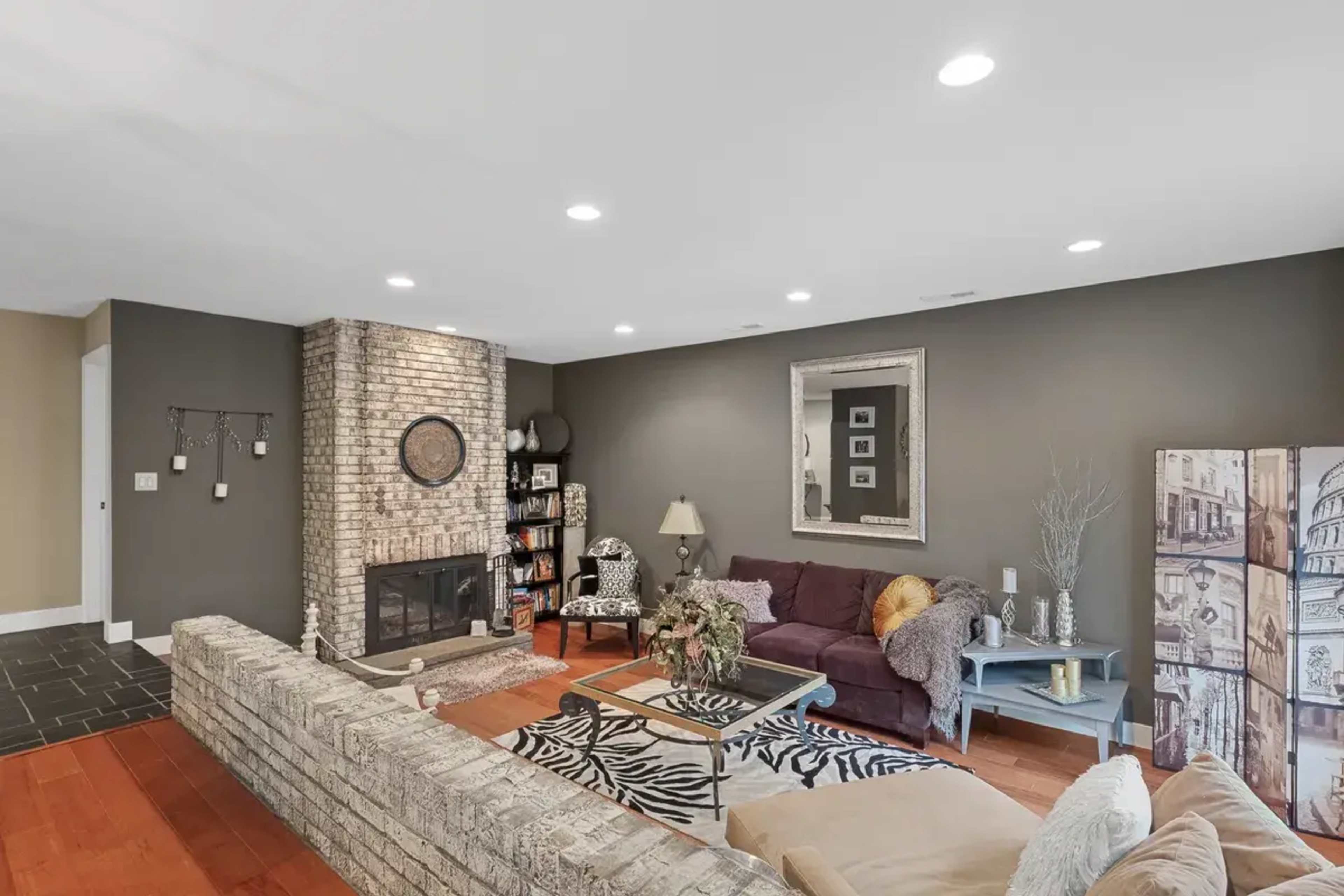 The living room features a brick fireplace, a sectional sofa, and a coffee table on an area rug, surrounded by dark walls and wooden flooring.