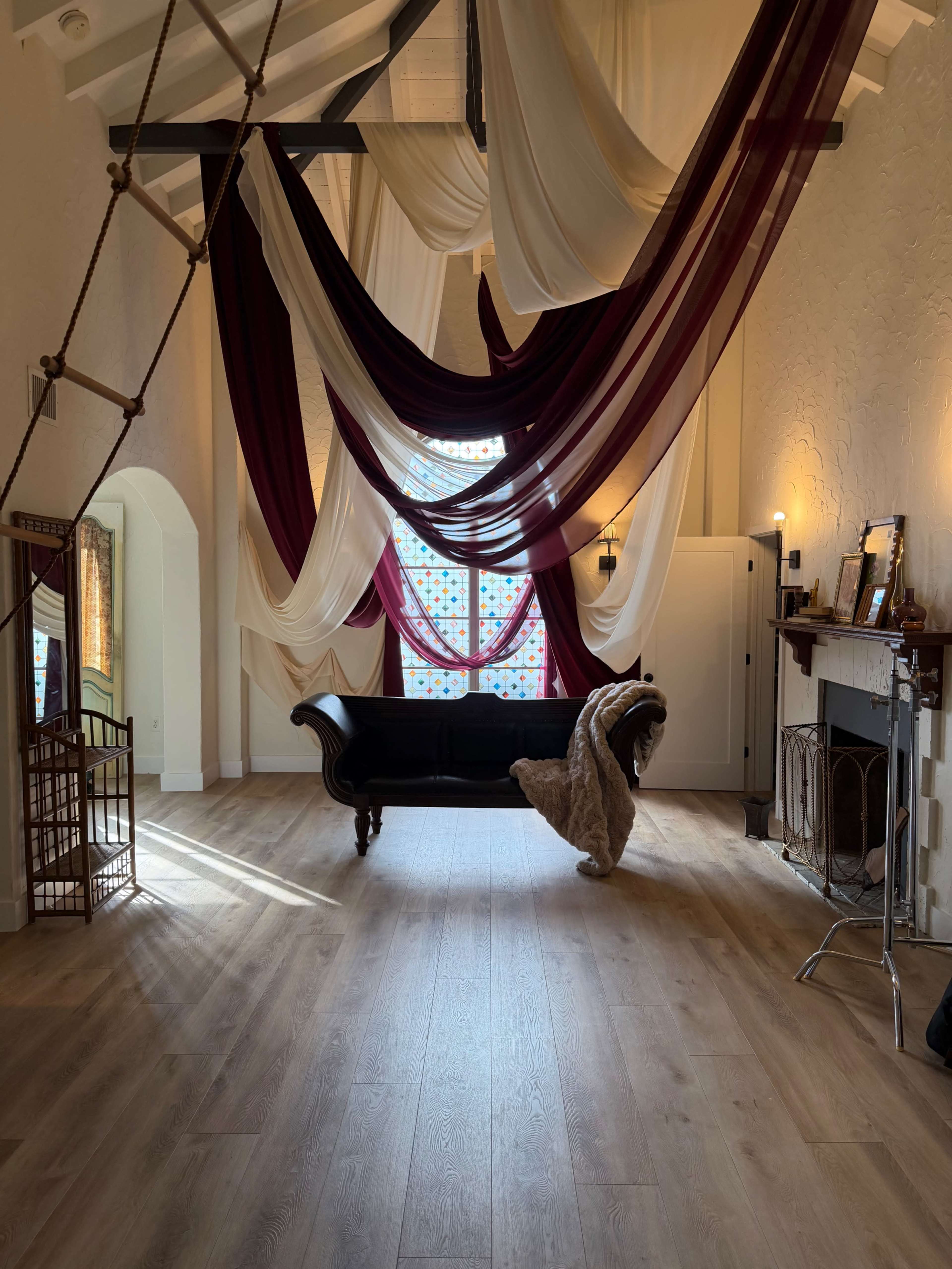 The image shows a spacious interior with draped fabric in maroon and beige hanging from the ceiling, a black couch in the center, and warm lighting creating a cozy ambiance.