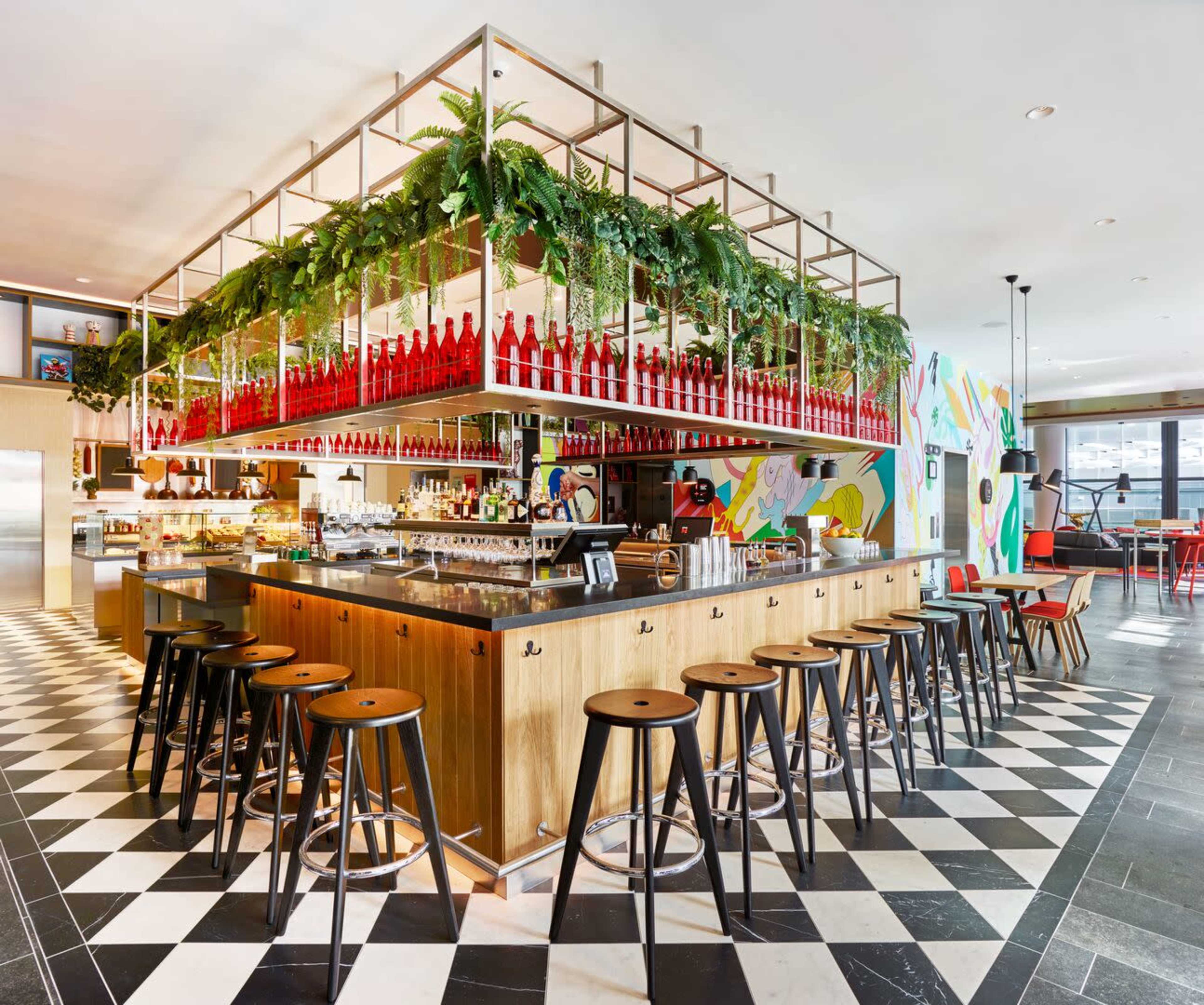 A modern bar area featuring a central counter surrounded by stools, adorned with hanging plants and rows of bottles above.