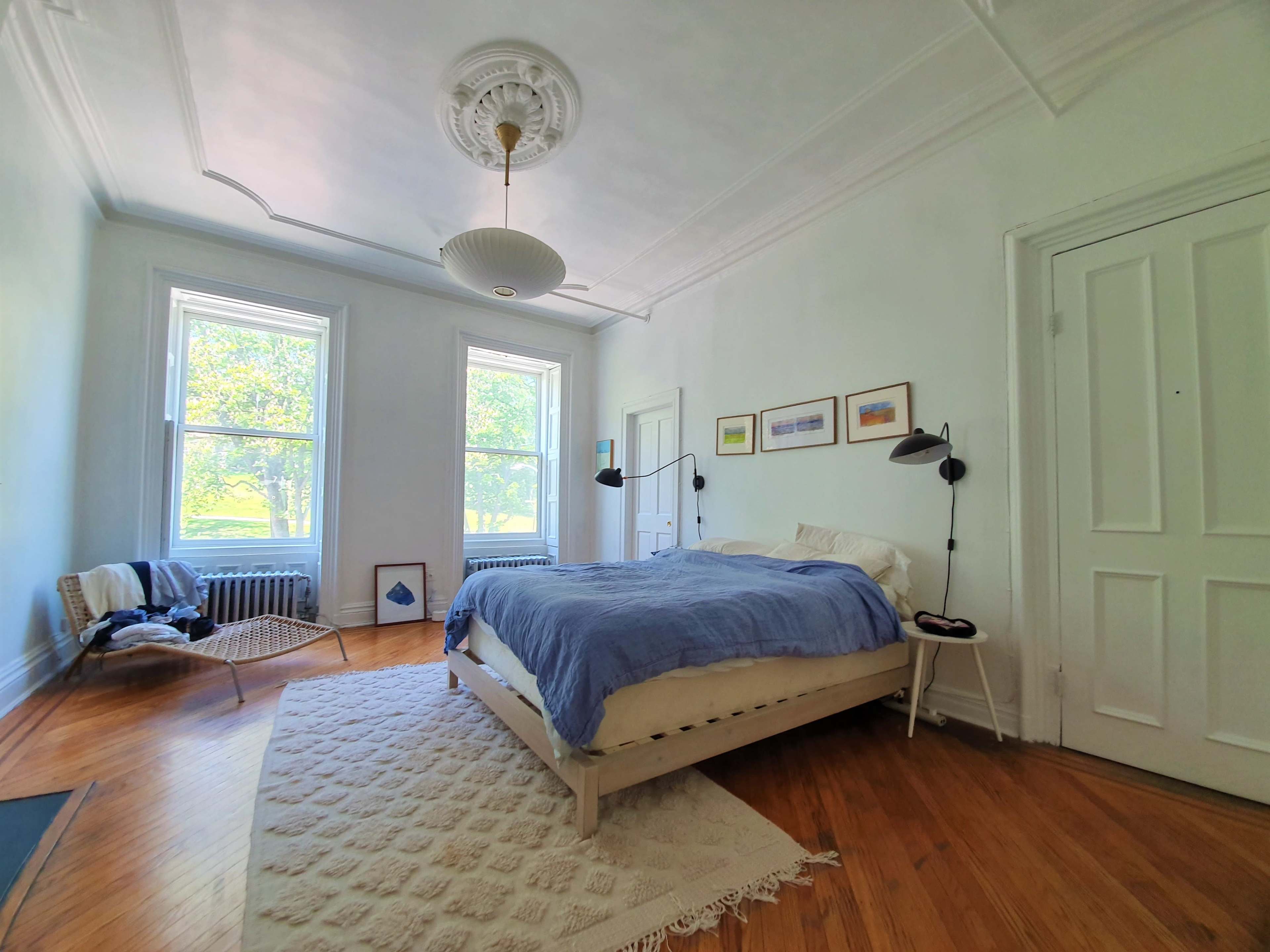 A bright, airy bedroom with large windows, a bed with a blue blanket, and a chair near the wall.