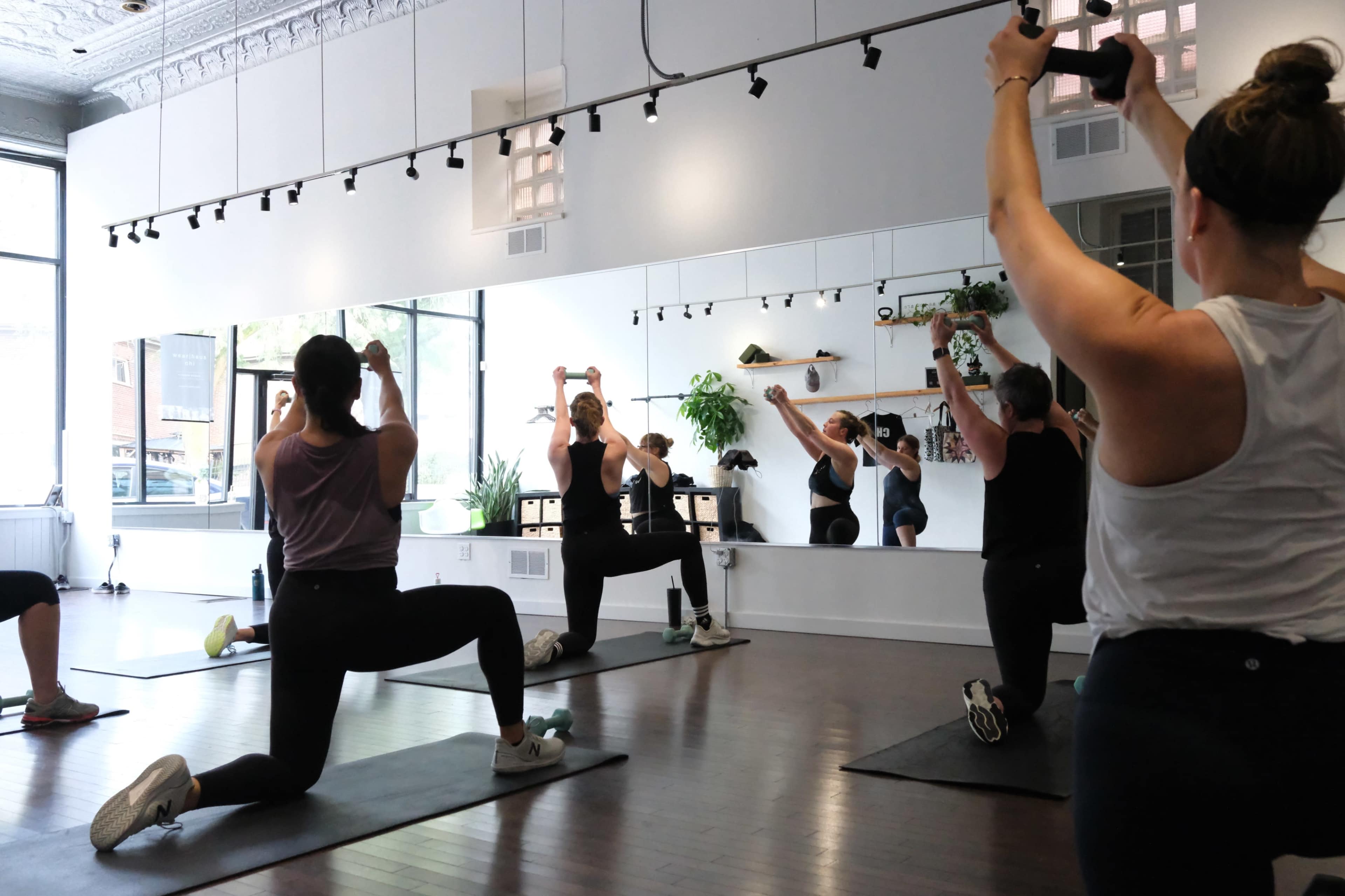 A group of women is engaged in a workout session in a fitness studio, using dumbbells and performing various exercises in front of a mirror.
