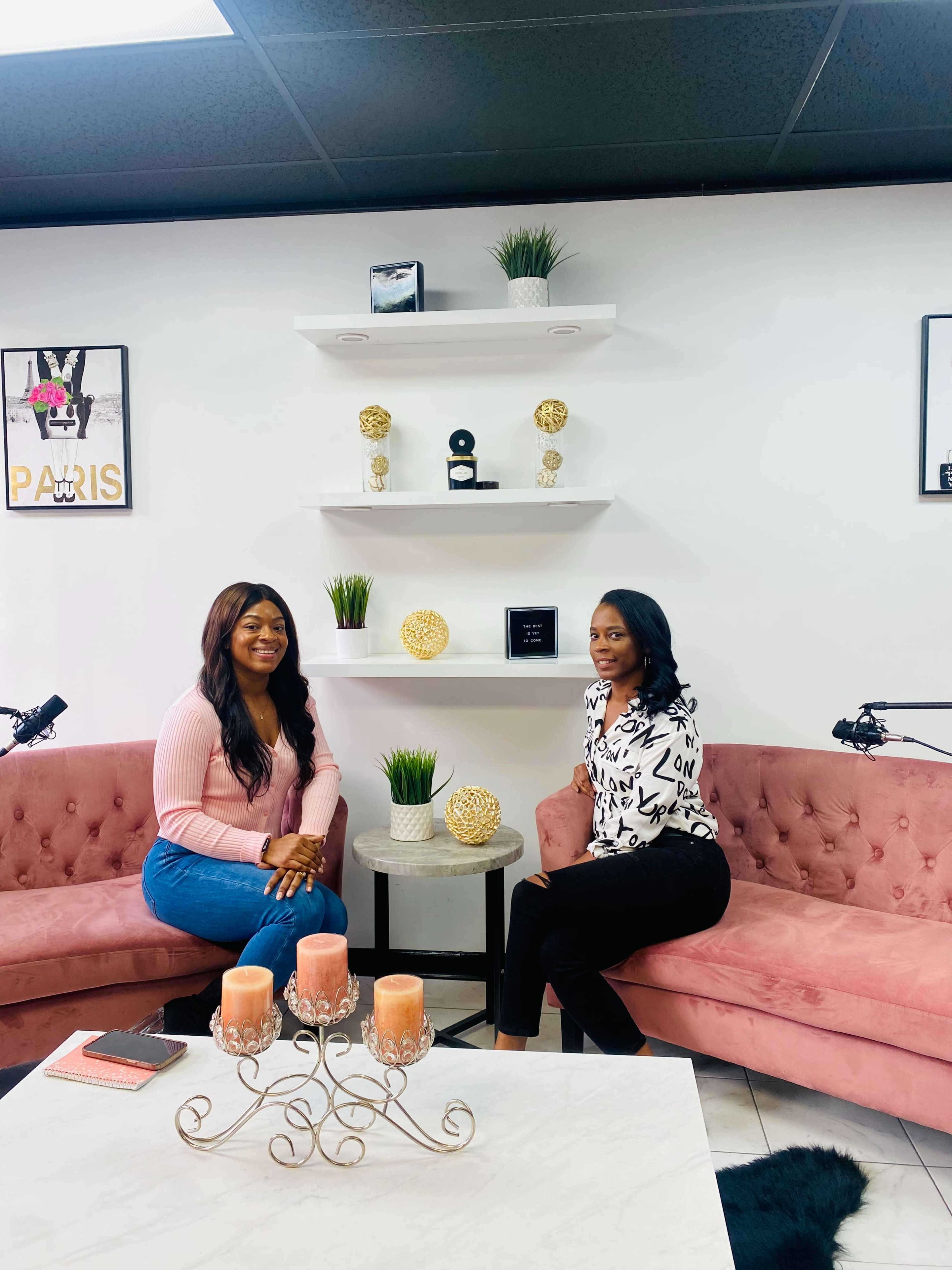 Two women sit on pink couches in a modern, stylish room with a white decorative shelf and candles on a coffee table.