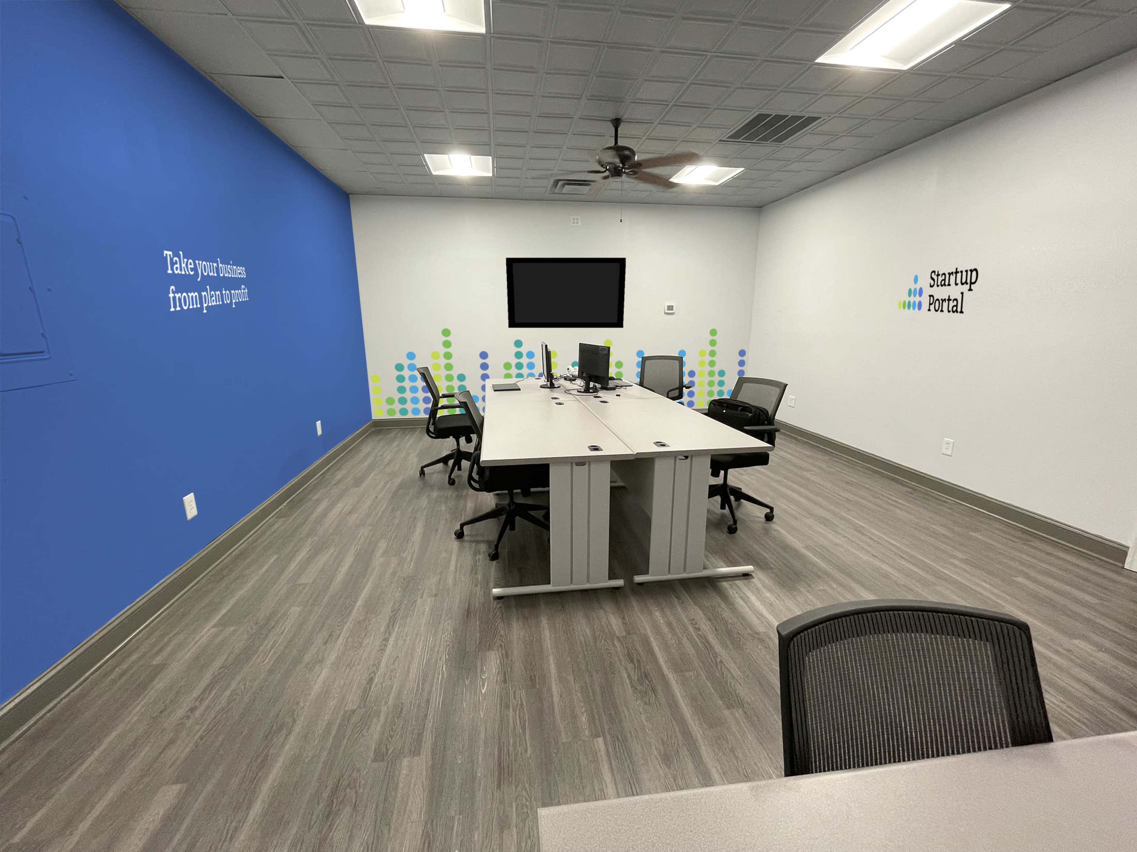A modern, empty meeting room with a large table, four chairs, a wall-mounted television, and motivational wall graphics.