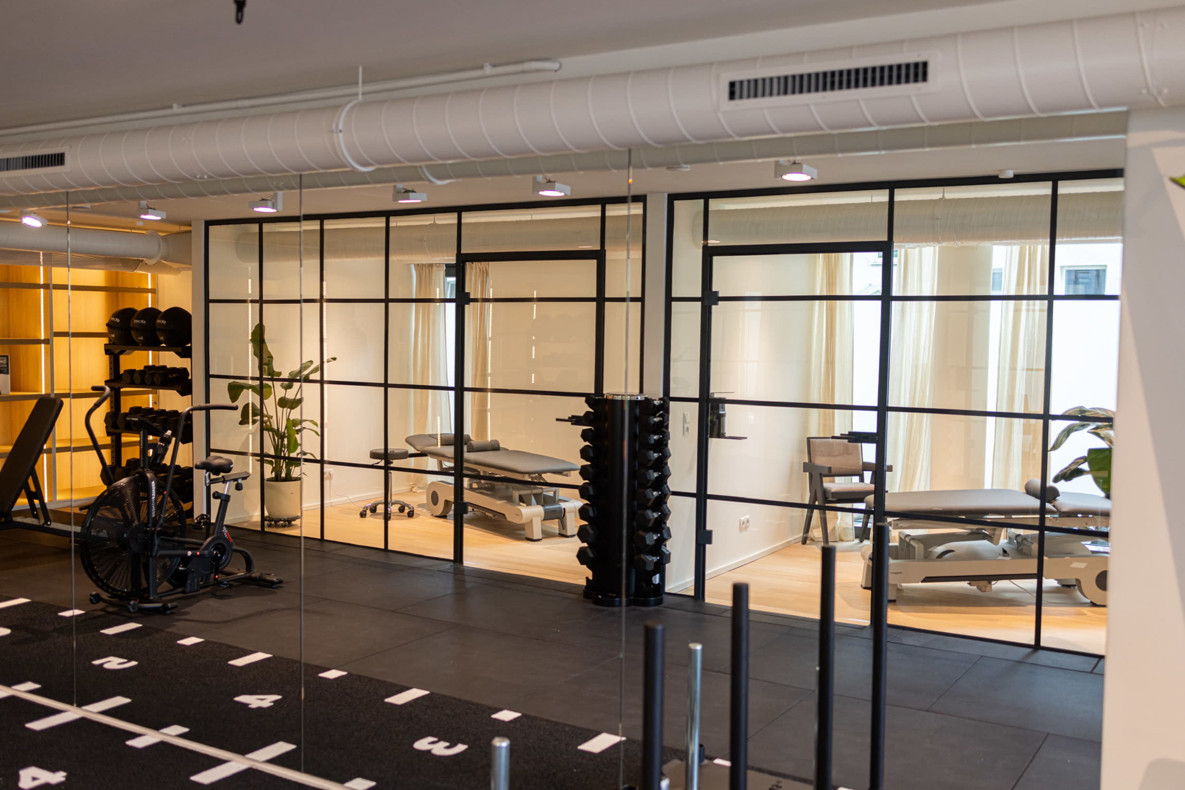 The image shows a modern gym space with black rubber flooring, fitness equipment on one side, and a glass-walled area featuring exercise tables and greenery visible through the windows.