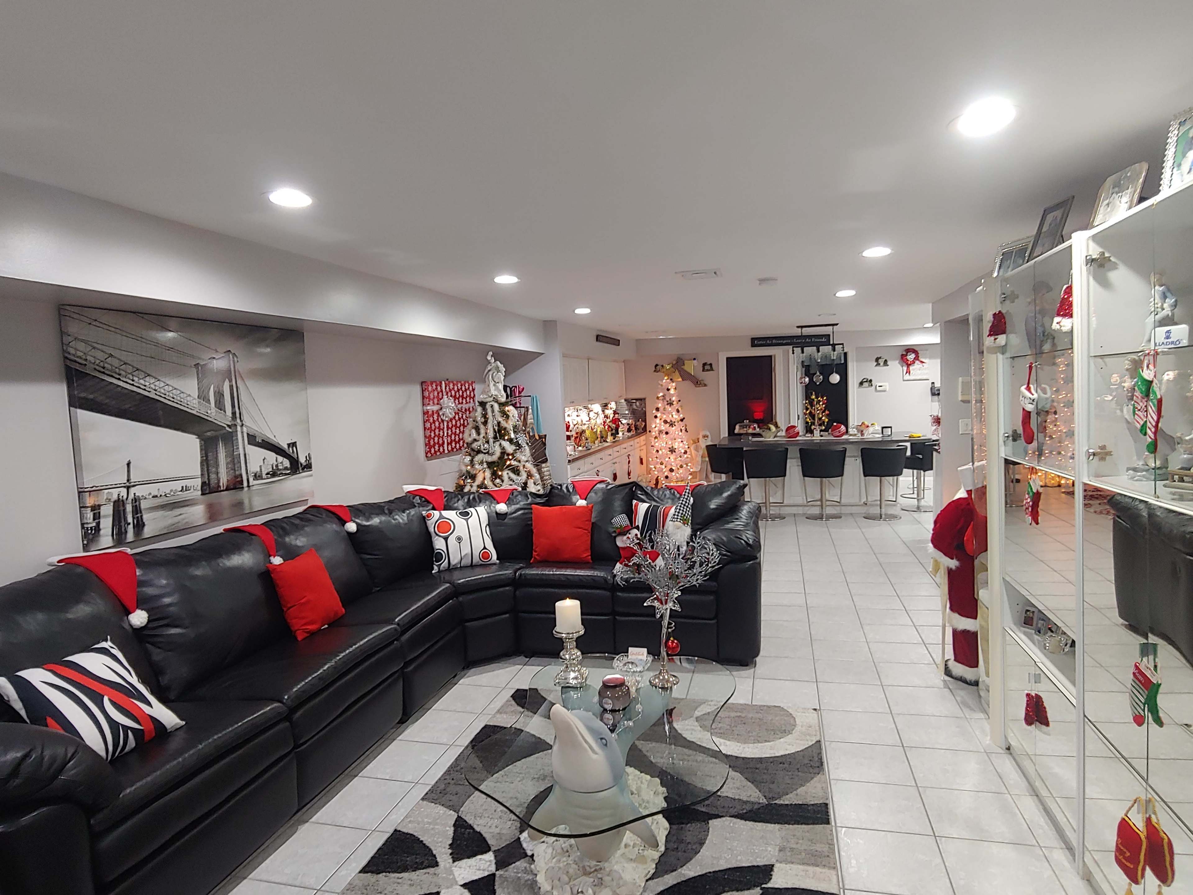 A modern living room decorated for the holidays, featuring a black sectional sofa with red and white cushions, a glass coffee table, and a Christmas tree in the corner.