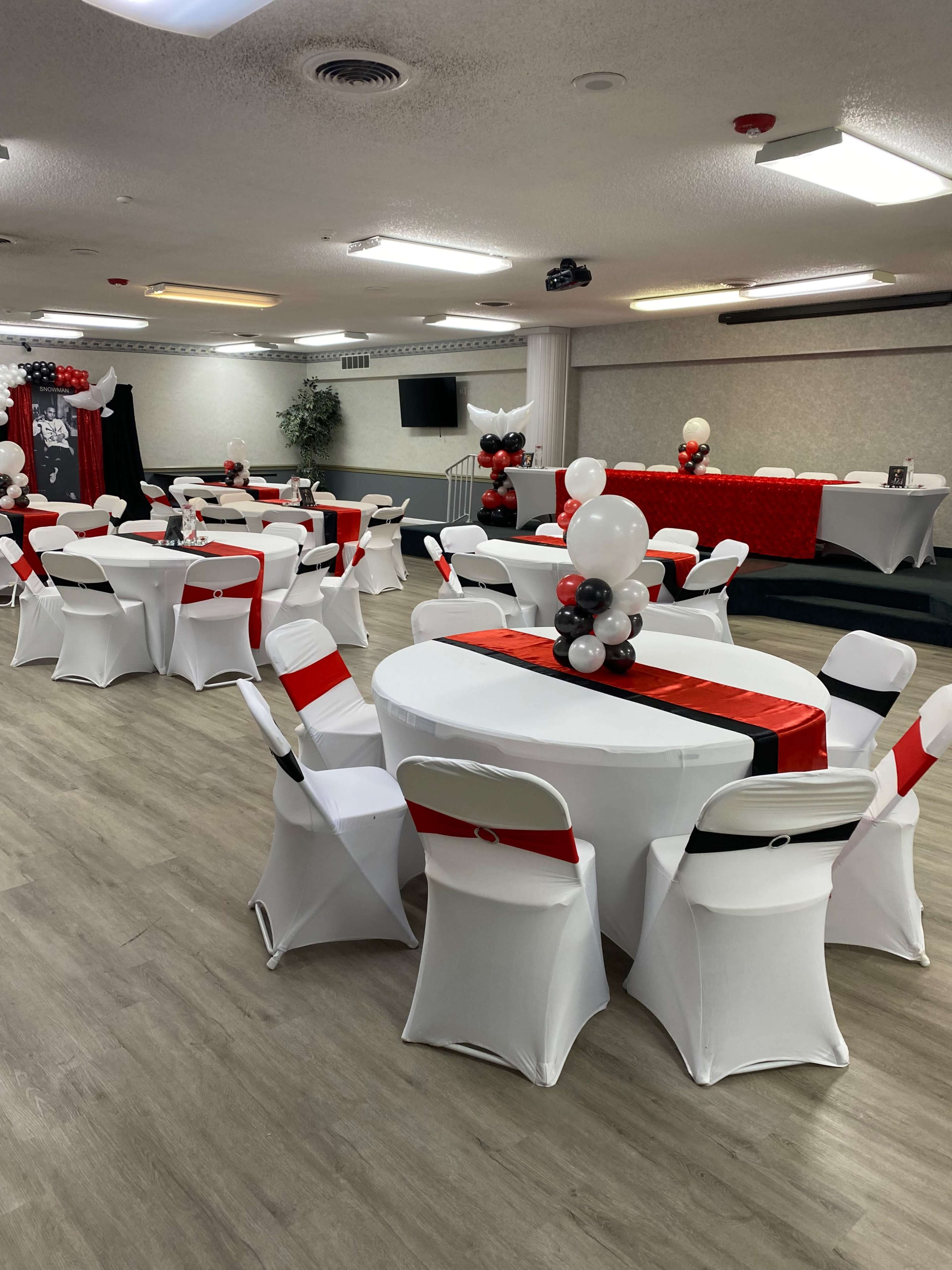 A decorated event space featuring white tables with red accents, white chairs, and balloon arrangements in black and white.