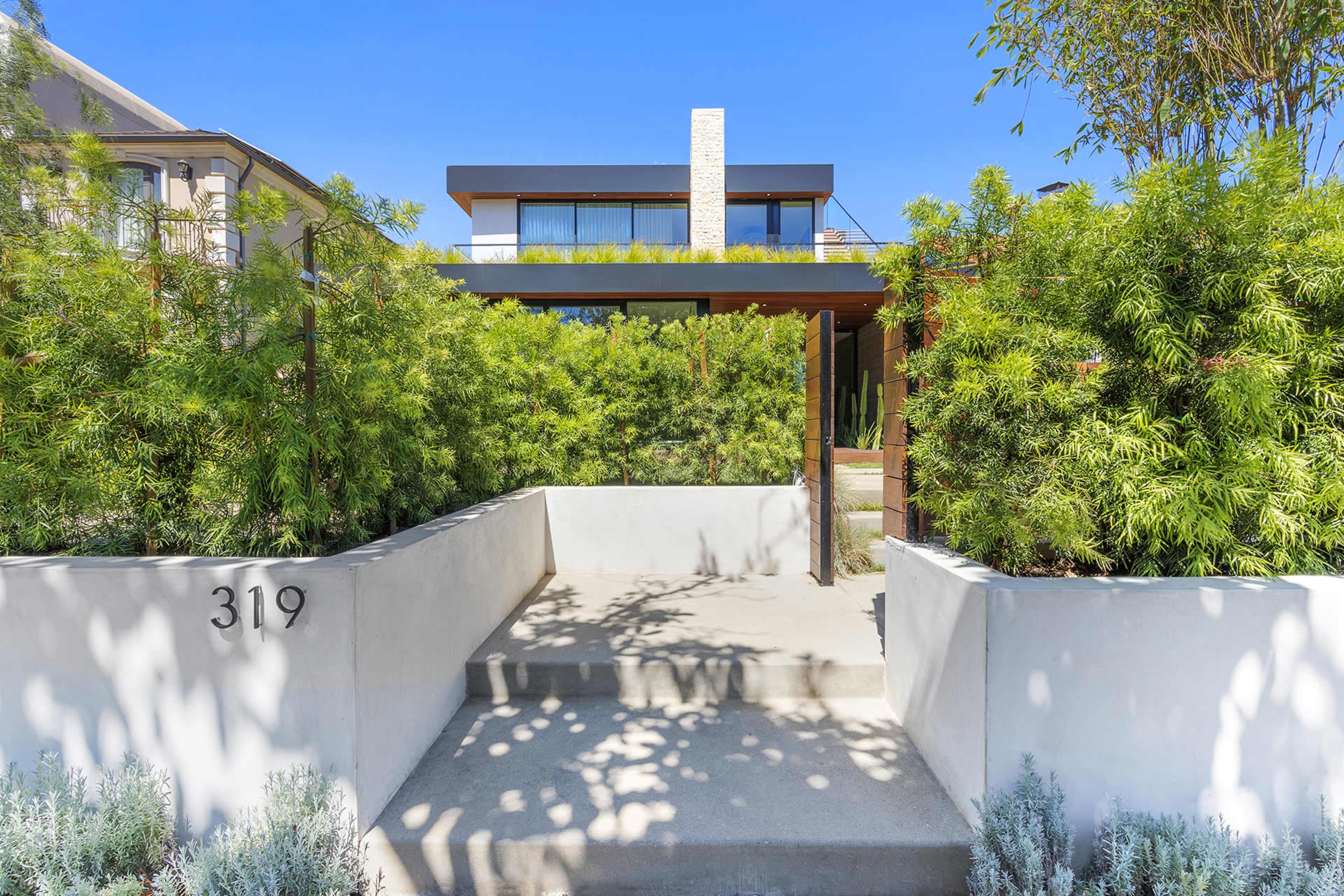 A modern home with a stone chimney is surrounded by lush greenery and features a concrete pathway leading to the entrance.