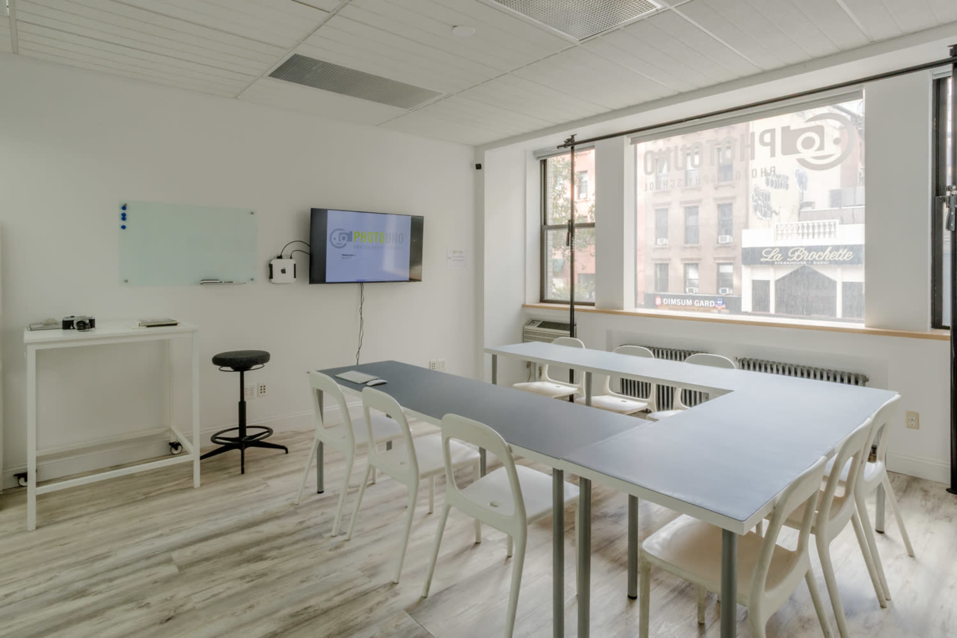 The image shows a bright, modern classroom with a long table, white chairs, a TV screen on the wall, and large windows letting in natural light.