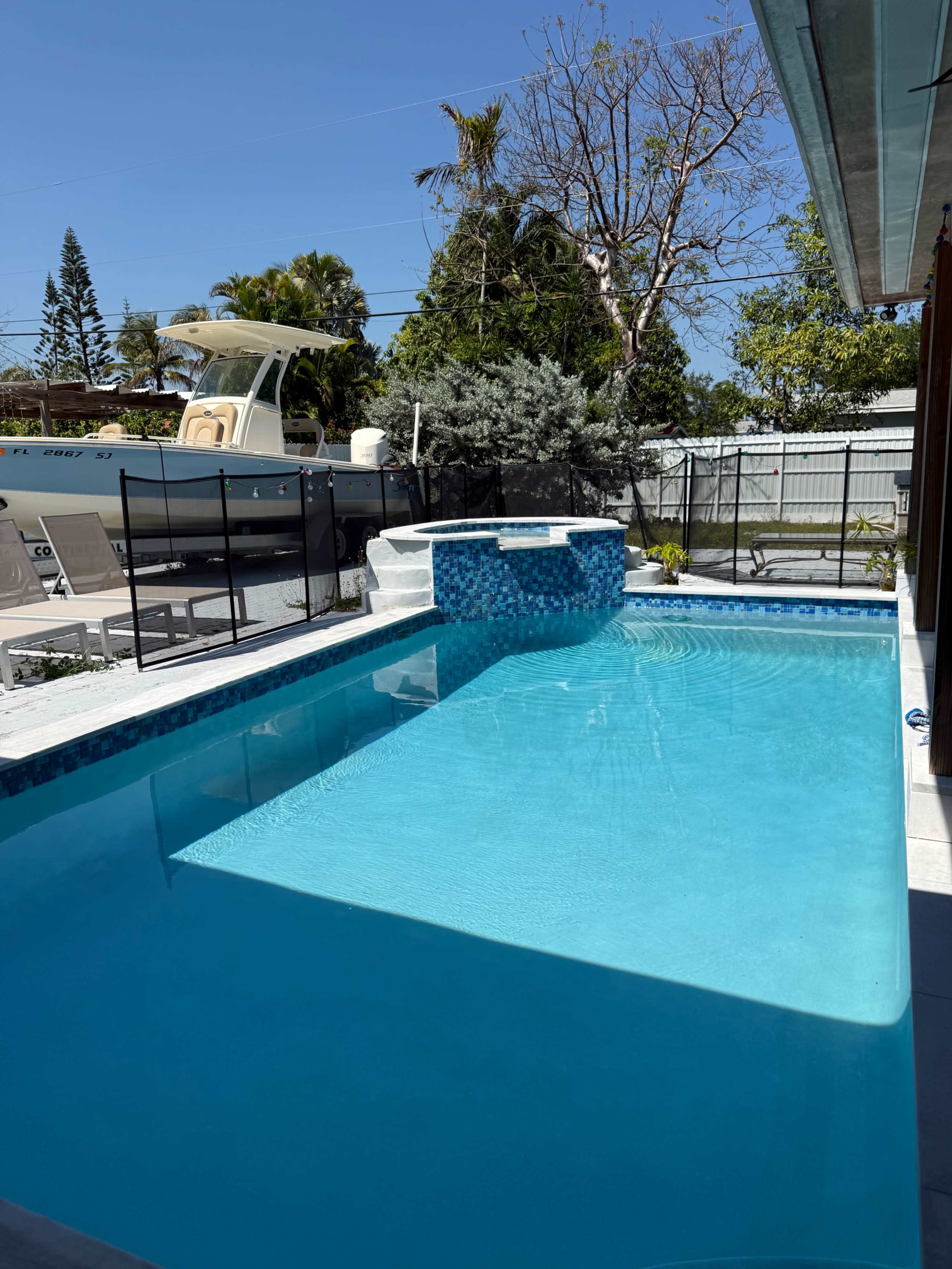 The image shows a clear swimming pool surrounded by a patio and vegetation, with a boat parked nearby under a bright blue sky.
