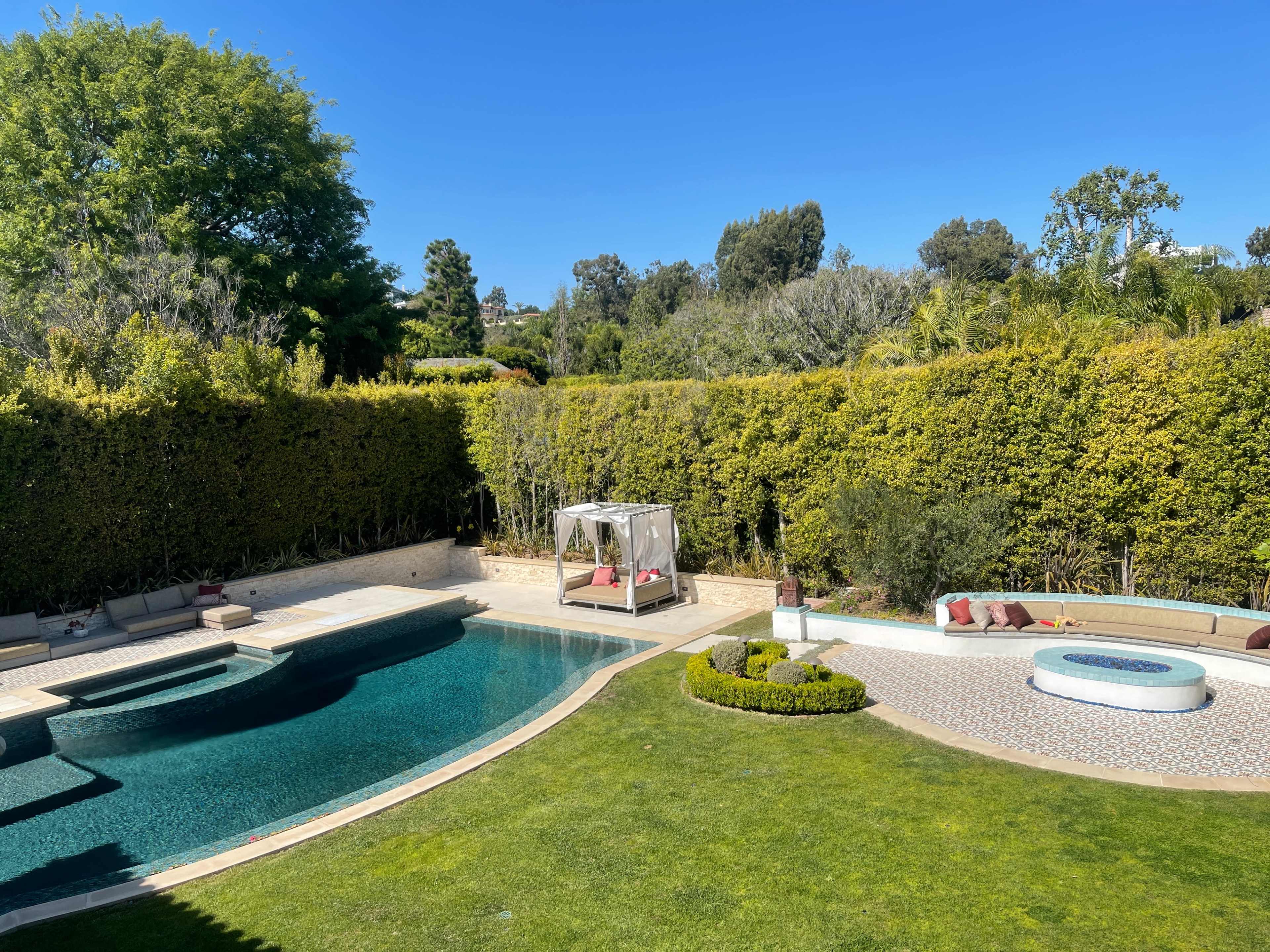 A well-maintained backyard features a swimming pool, a seating area, and a gazebo surrounded by lush greenery and a clear blue sky.