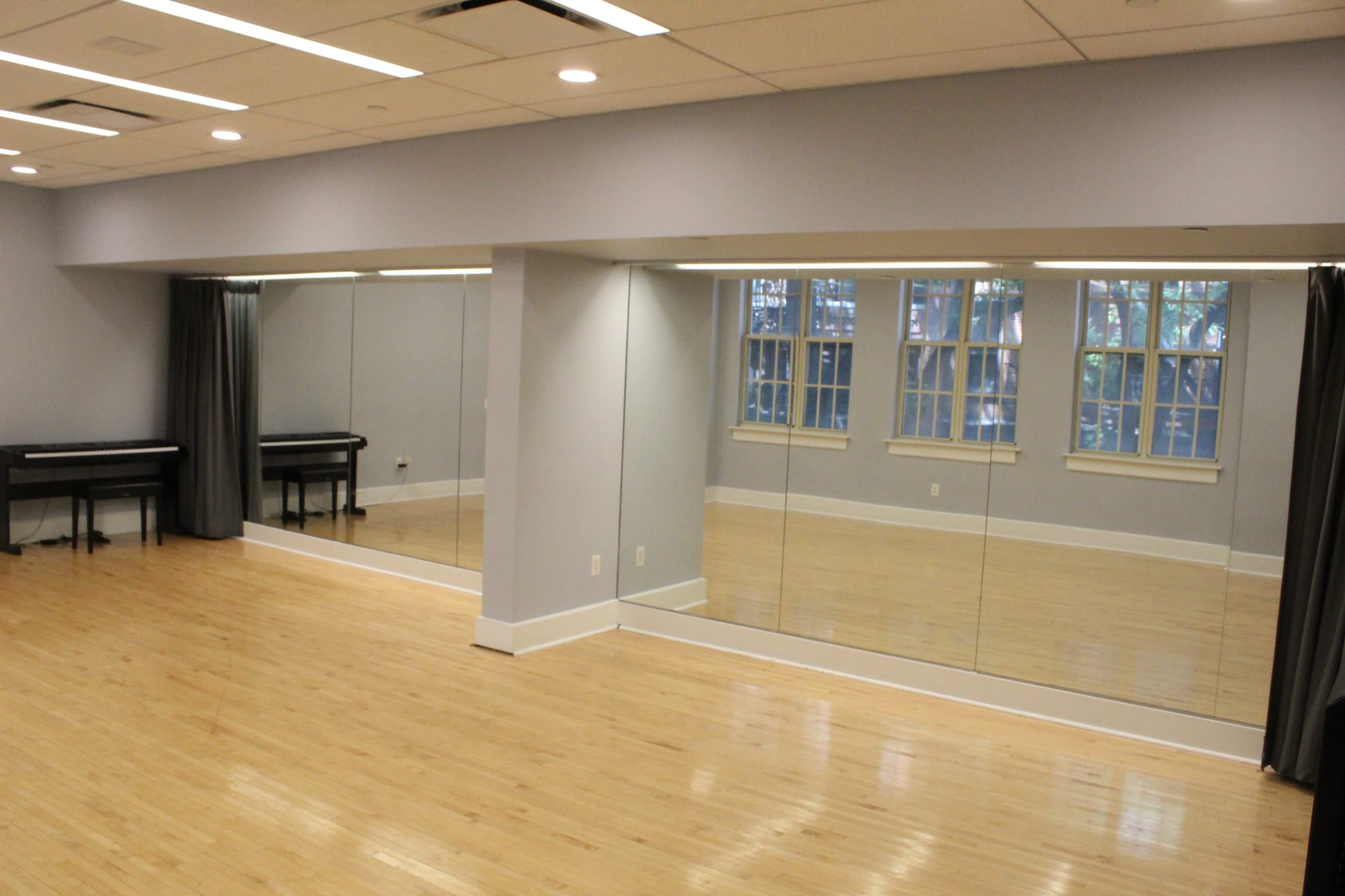 The image shows a spacious room with a polished wooden floor, a wall of mirrors, and a piano in the corner beneath large windows.