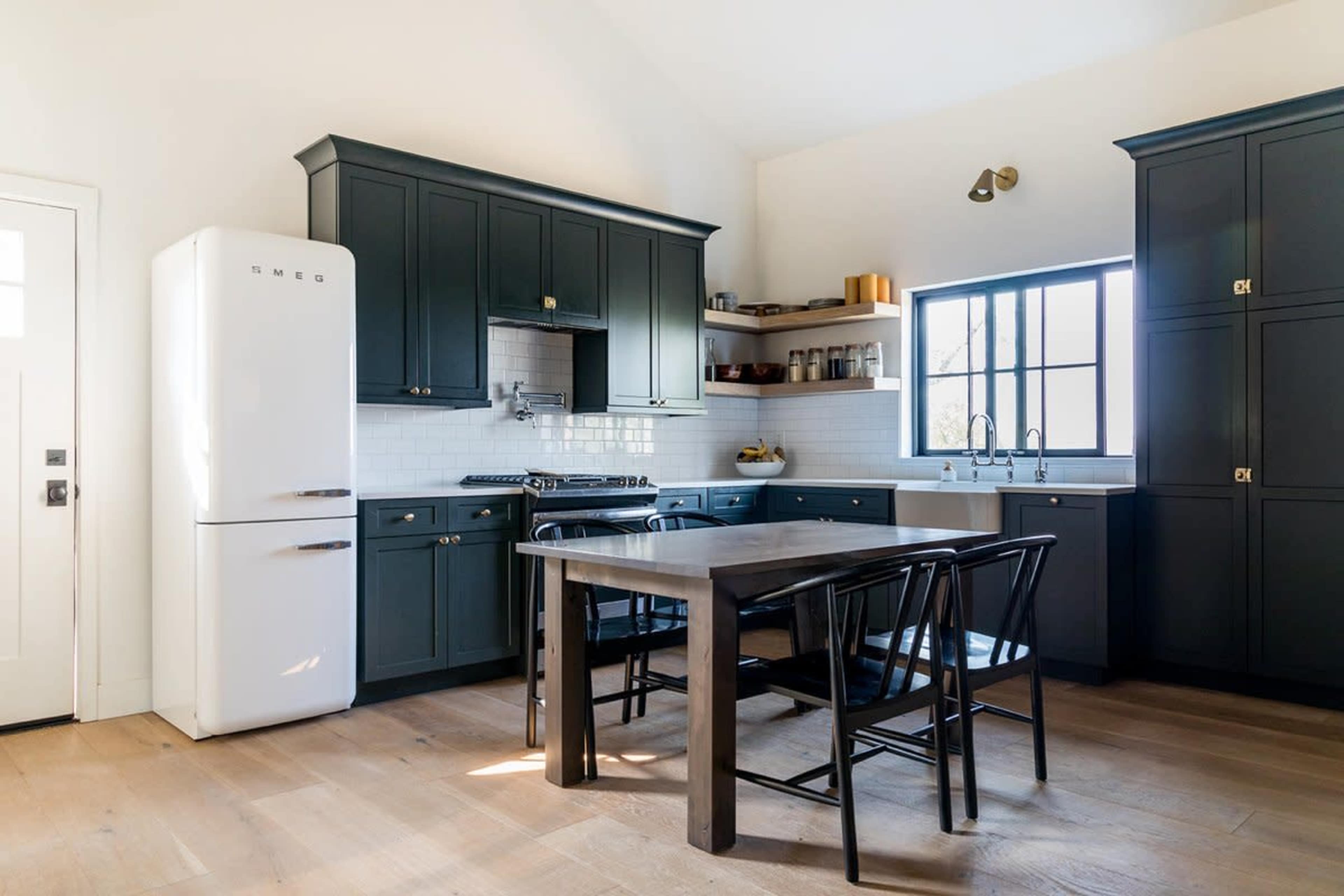 The image shows a modern kitchen with dark cabinets, a white refrigerator, a wooden dining table, and black chairs.
