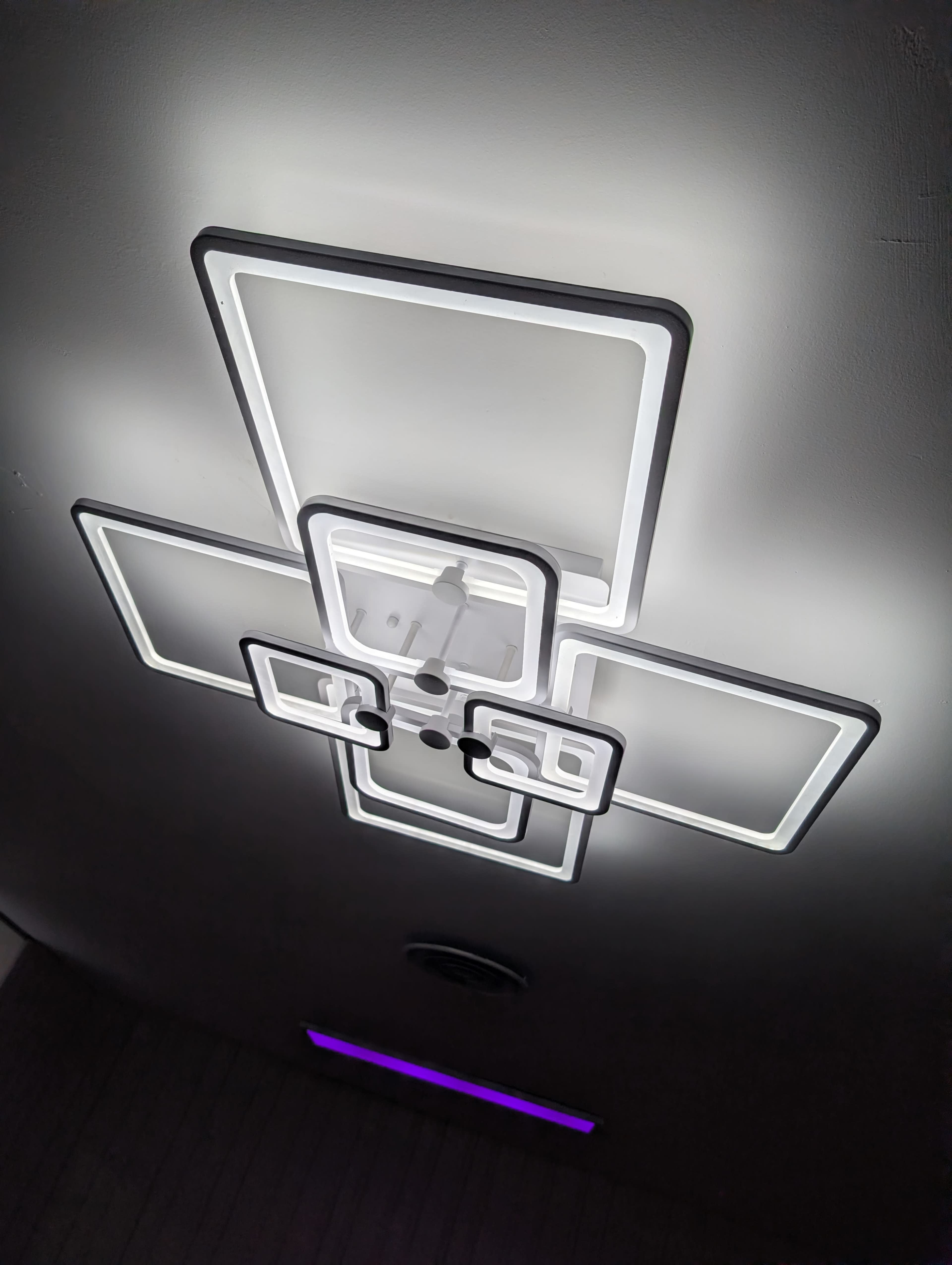 The image shows a modern ceiling light fixture composed of multiple interconnected rectangular shapes emitting a soft glow.
