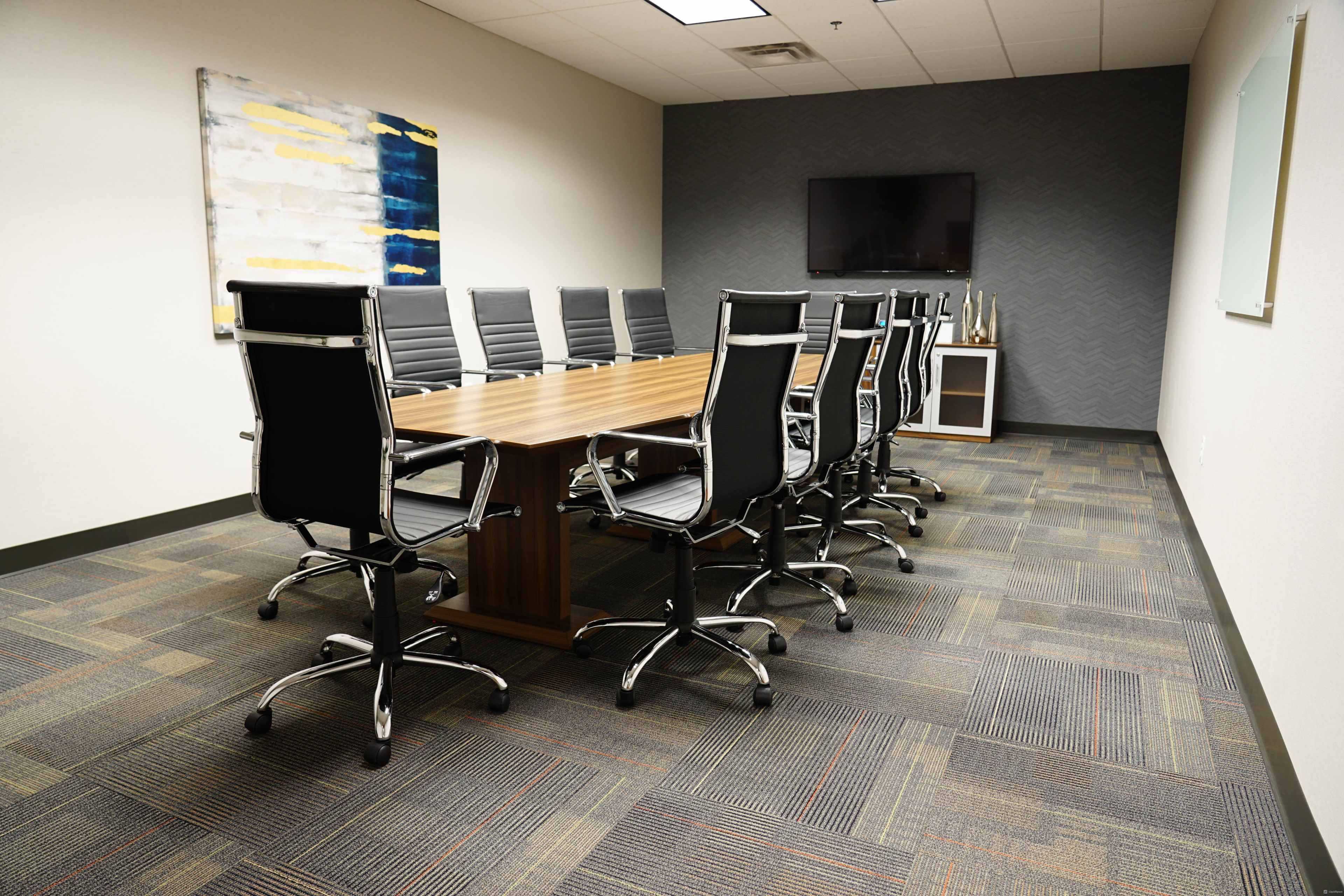 A conference room features a long wooden table surrounded by high-backed black chairs, with a television mounted on the wall and a whiteboard nearby.