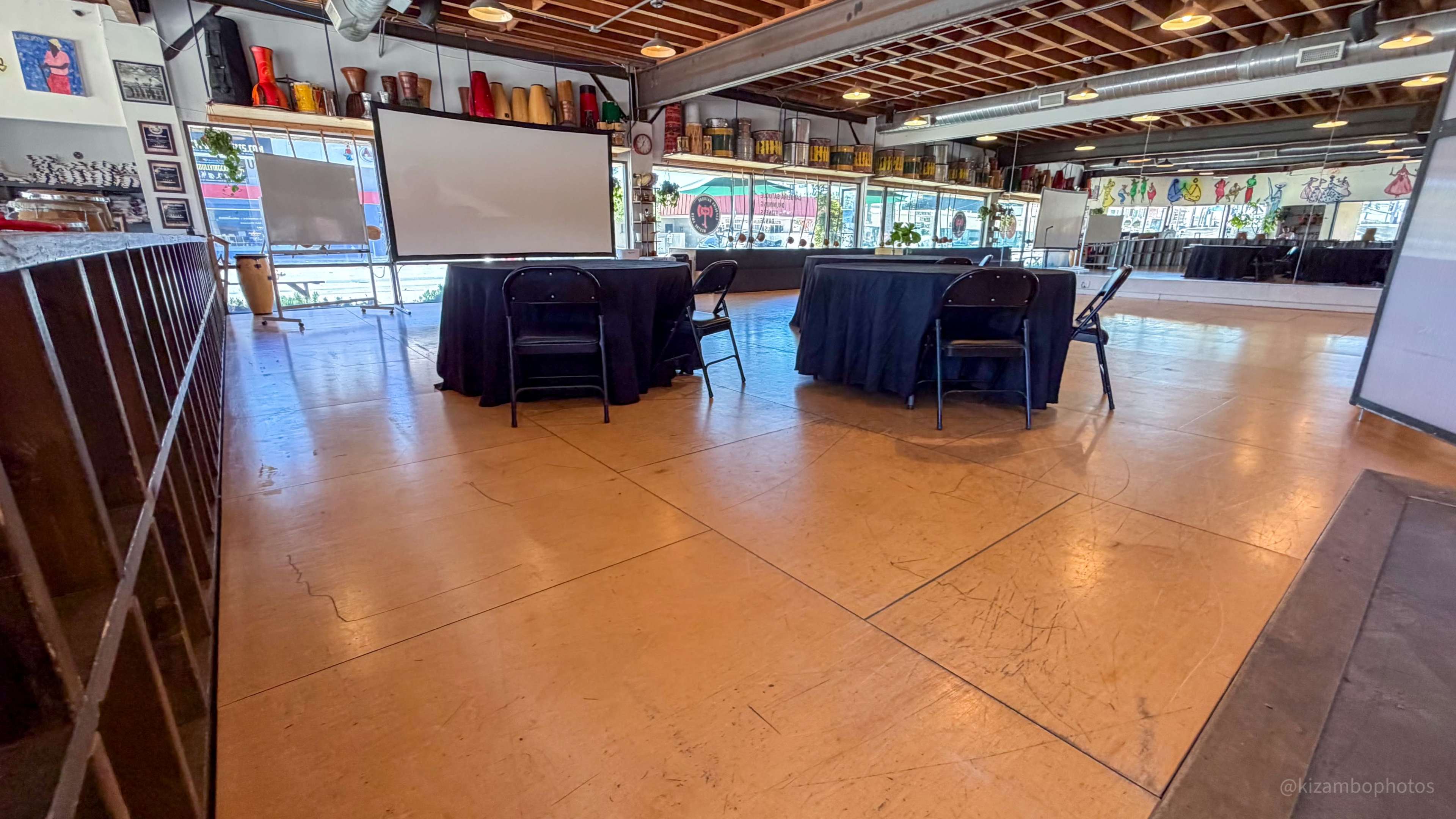 The image shows an interior space with a polished floor, several black tables with chairs, and a large projection screen in the background.