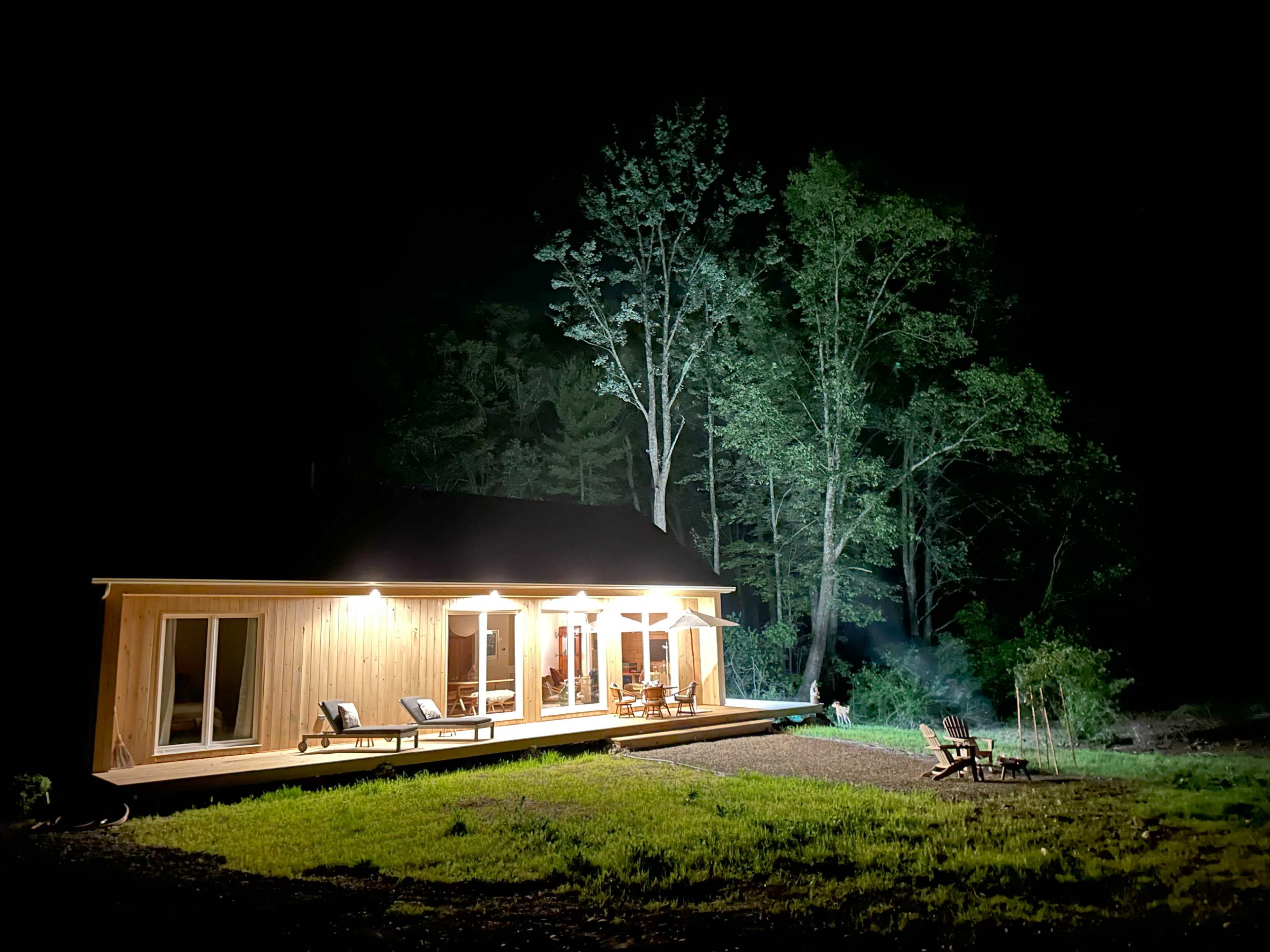 A wooden cabin is illuminated at night, surrounded by tall trees and a clear, dark sky.