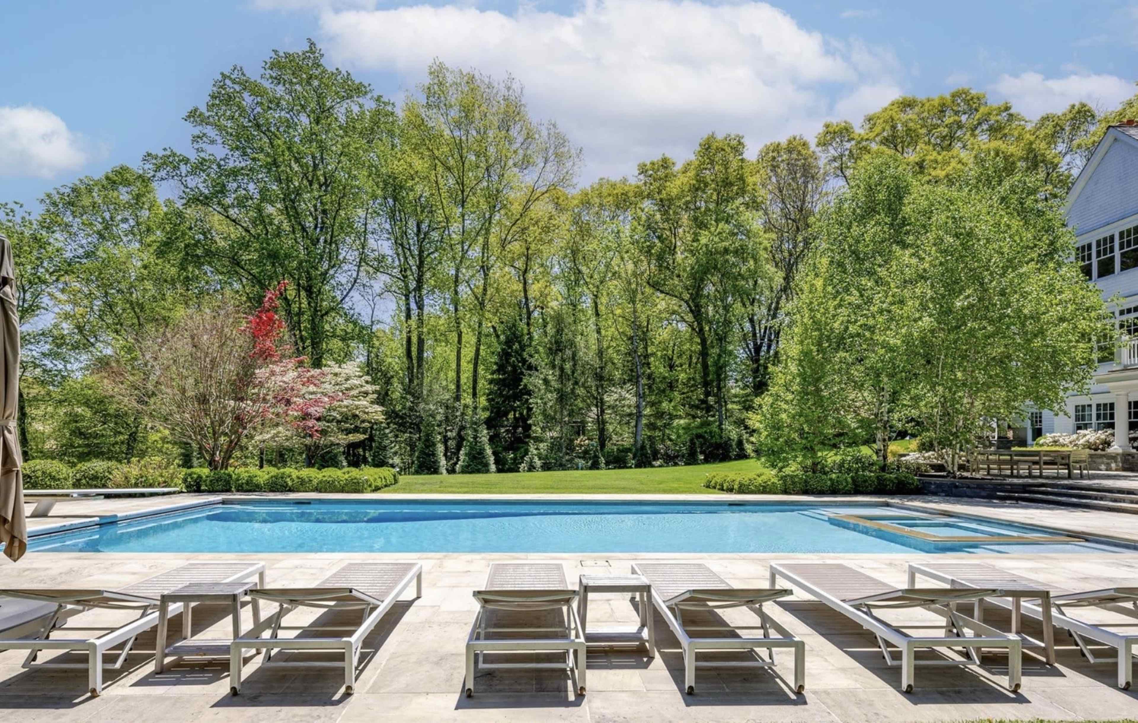 A large, residential swimming pool is surrounded by landscaped green lawns and trees on a sunny day.
