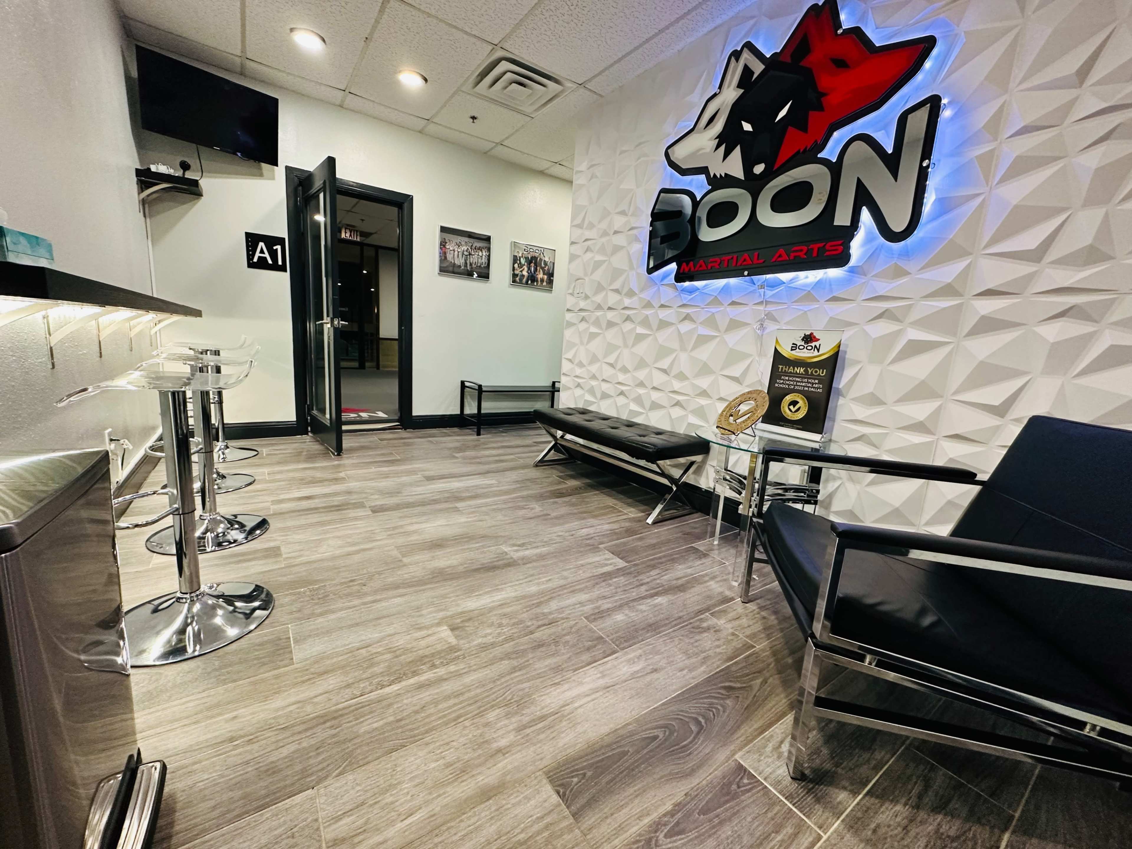 A modern waiting area for a martial arts studio, featuring sleek black furniture and a illuminated sign displaying the name "BOON MARTIAL ARTS."