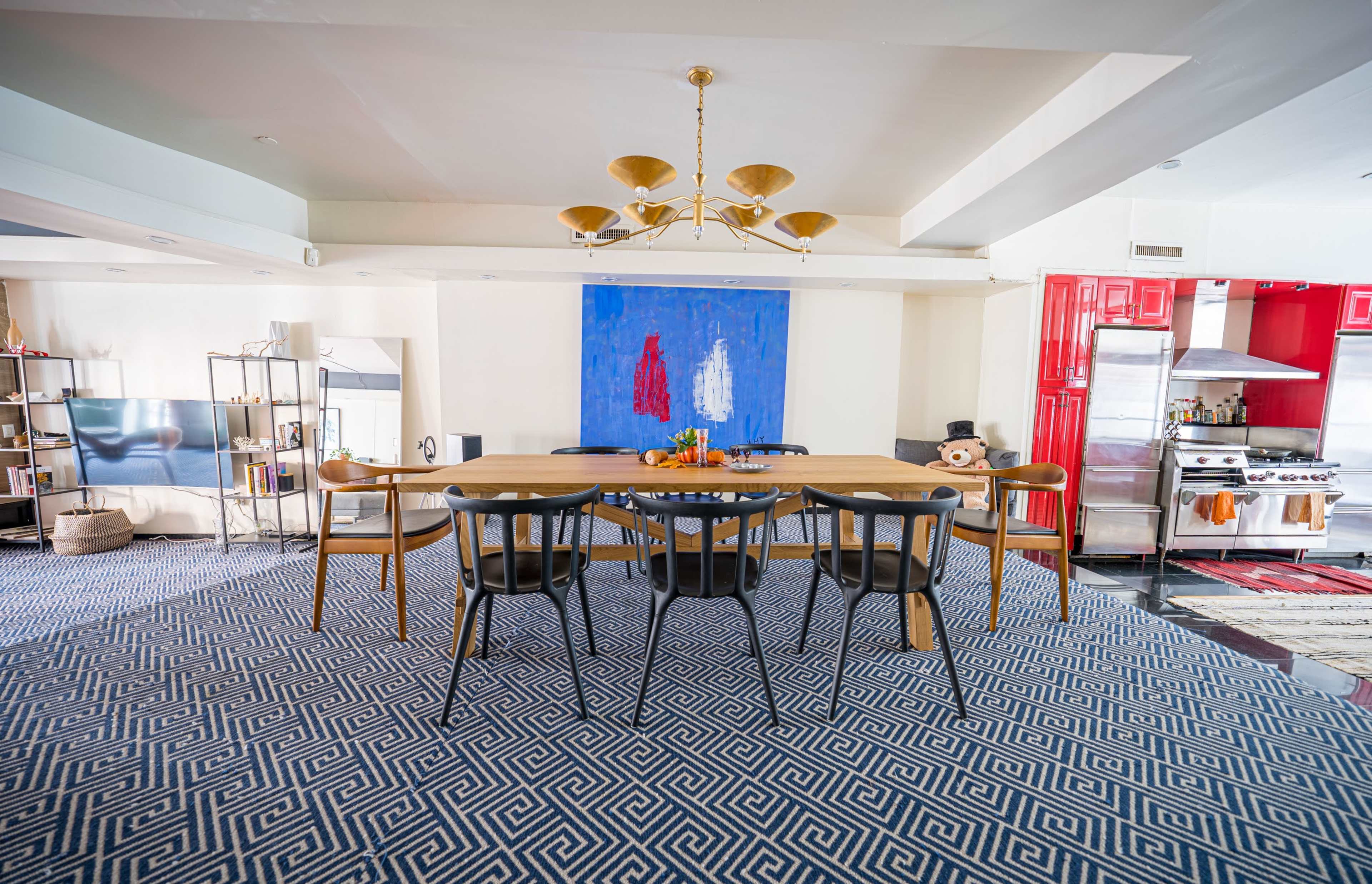 A modern dining area features a long wooden table surrounded by black chairs, with a blue abstract painting on the wall and a kitchen visible in the background.