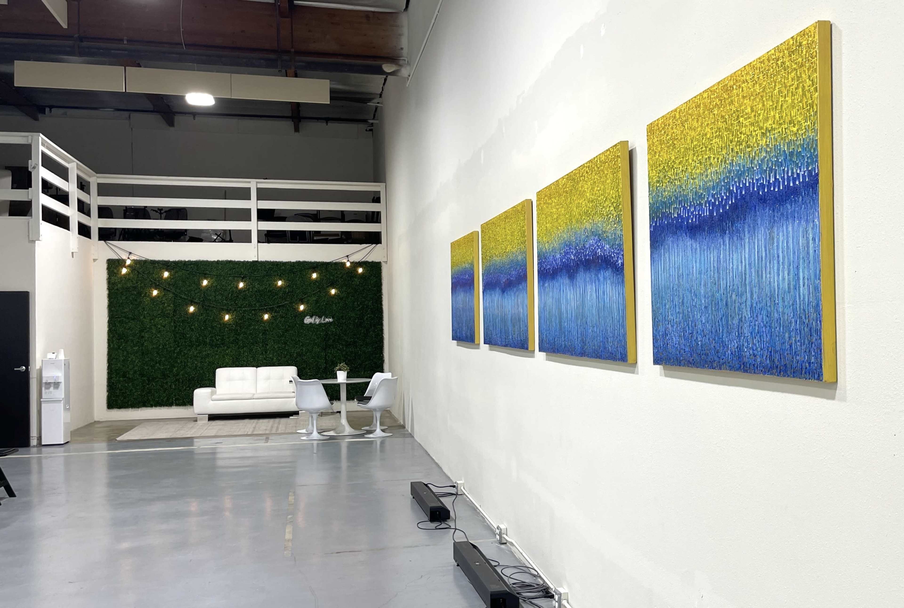 A minimalistic room featuring four colorful paintings on a white wall, a couch with chairs, and decorative string lights above a green wall.