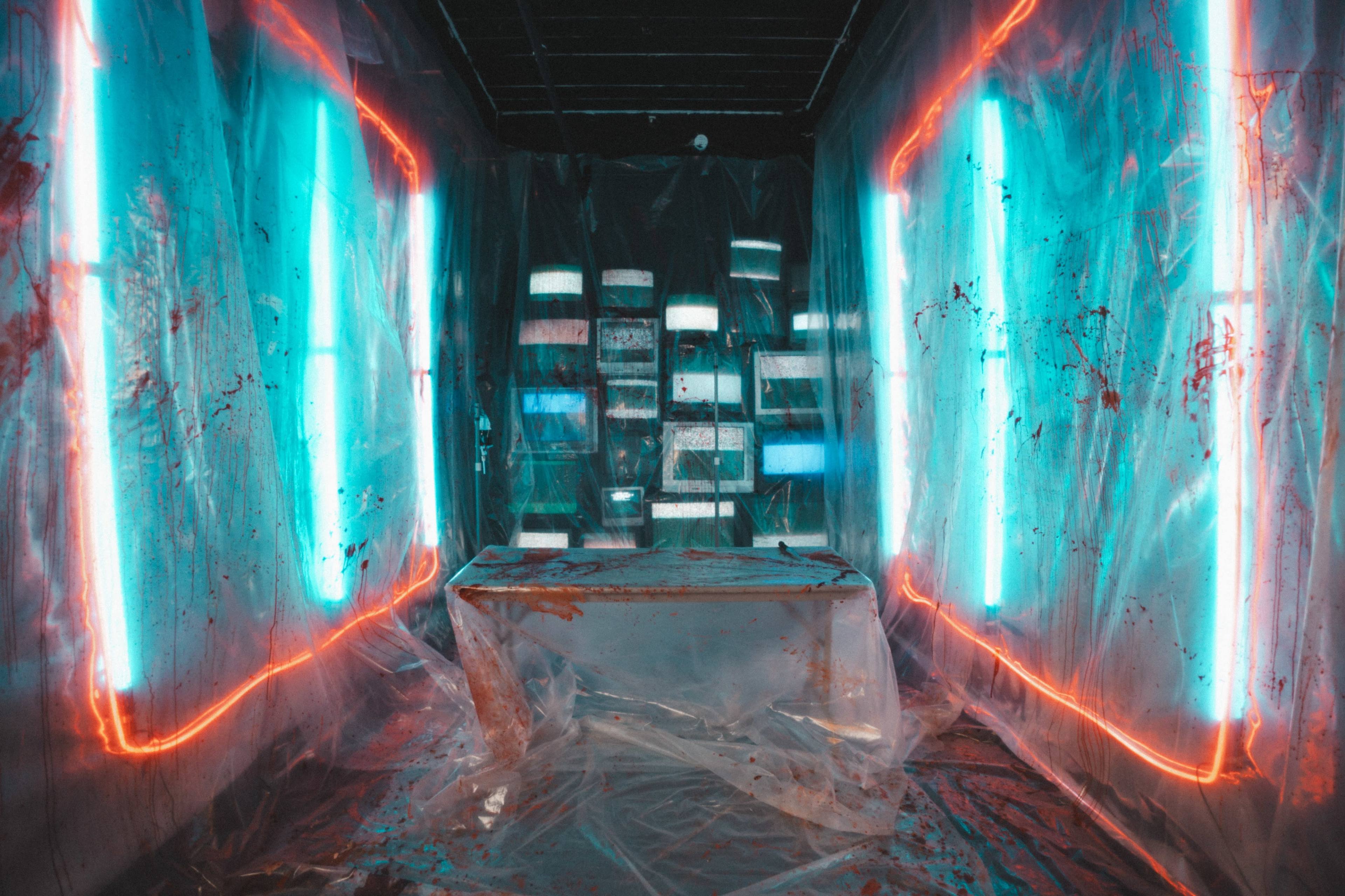 The image shows a dimly lit room with plastic sheeting covering the walls and a table in the center, illuminated by blue and red neon lights.