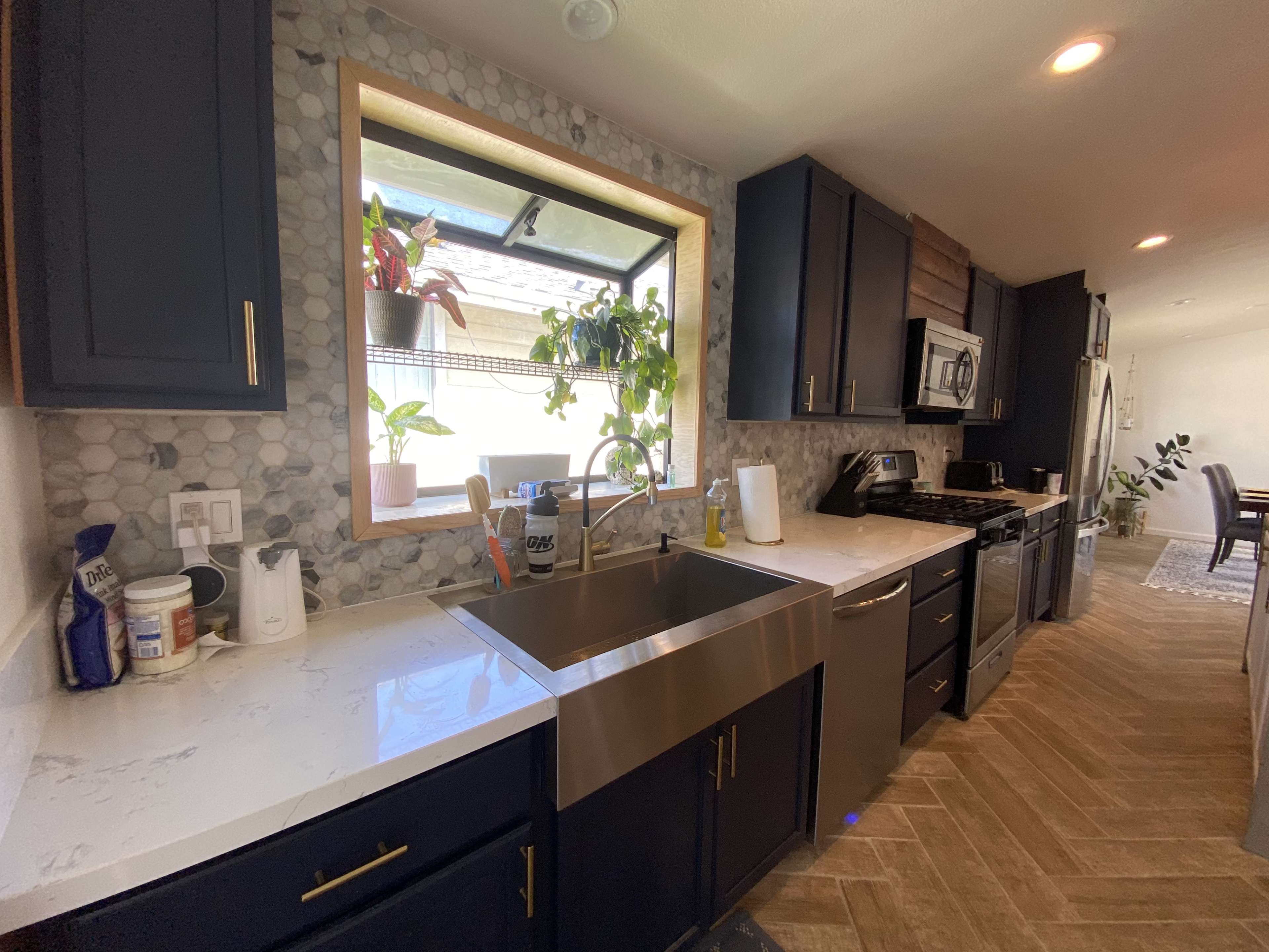 A modern kitchen with dark cabinets, a stainless steel sink, and a window surrounded by pebble tile, featuring plants on the sill.