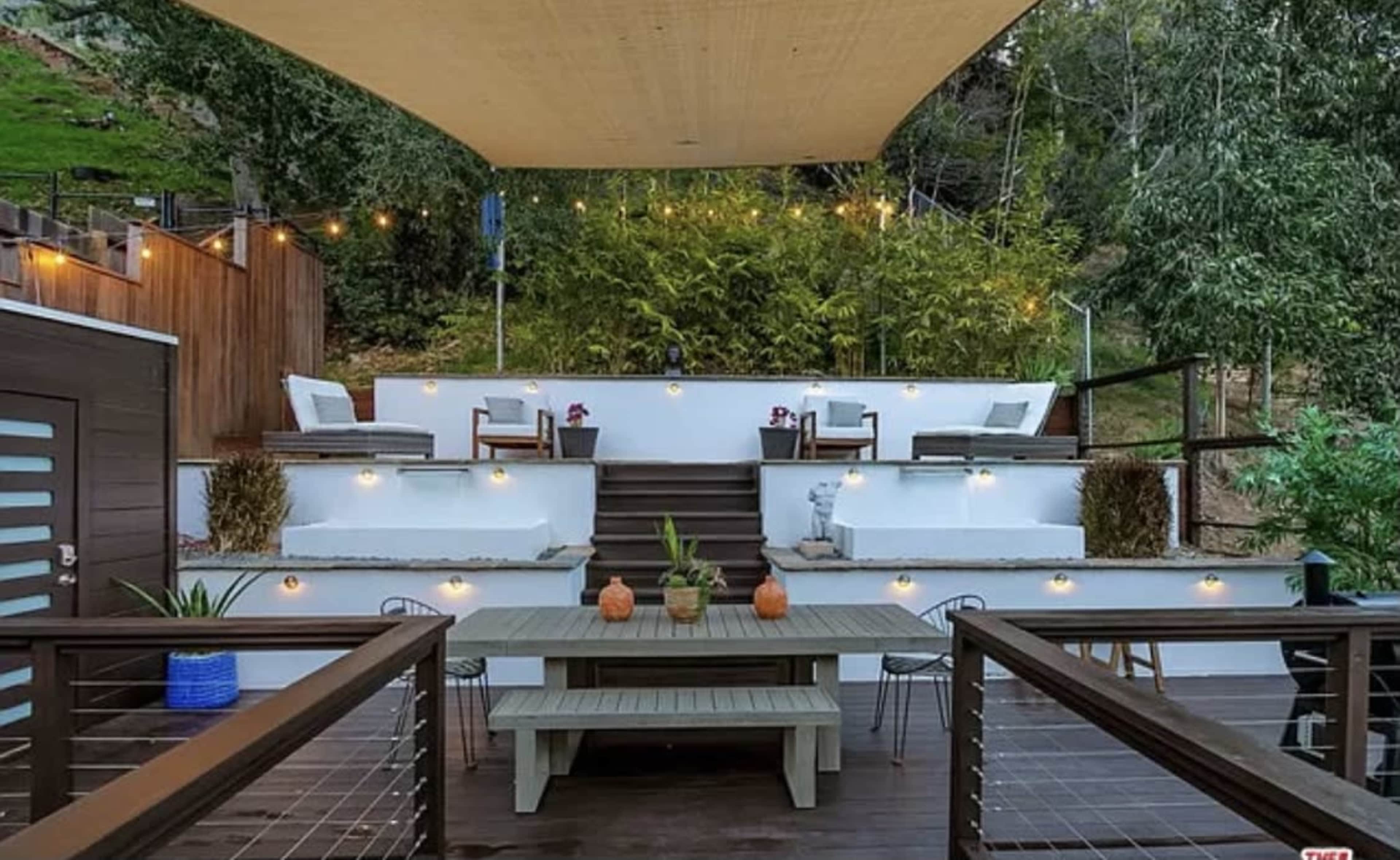 A modern outdoor patio area with a wooden dining table, decorative potted plants, and tiered seating areas surrounded by greenery and string lights.
