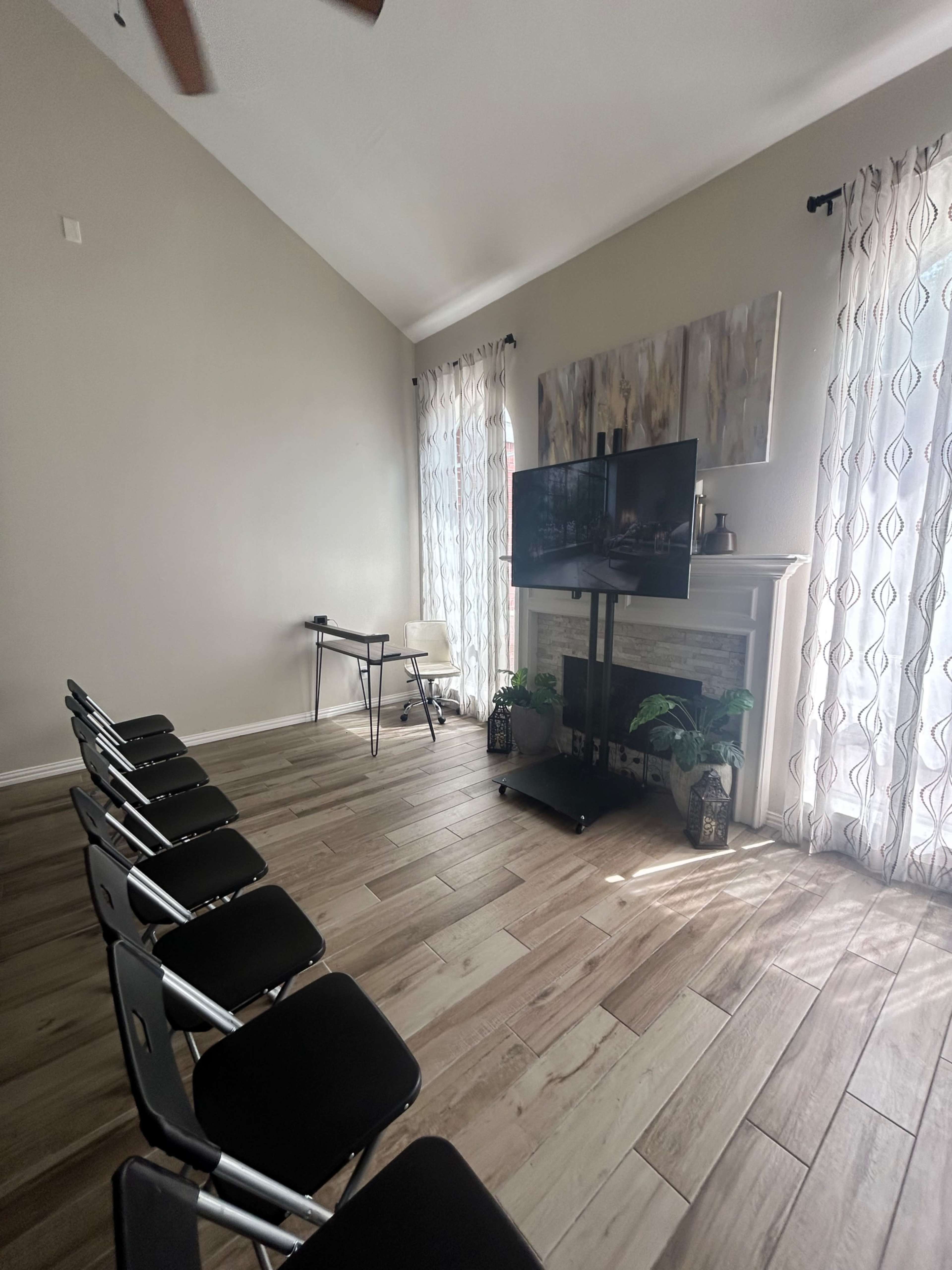 The image shows a room with light-colored walls, wooden flooring, a television mounted on a stand, and several folding chairs arranged in a row.