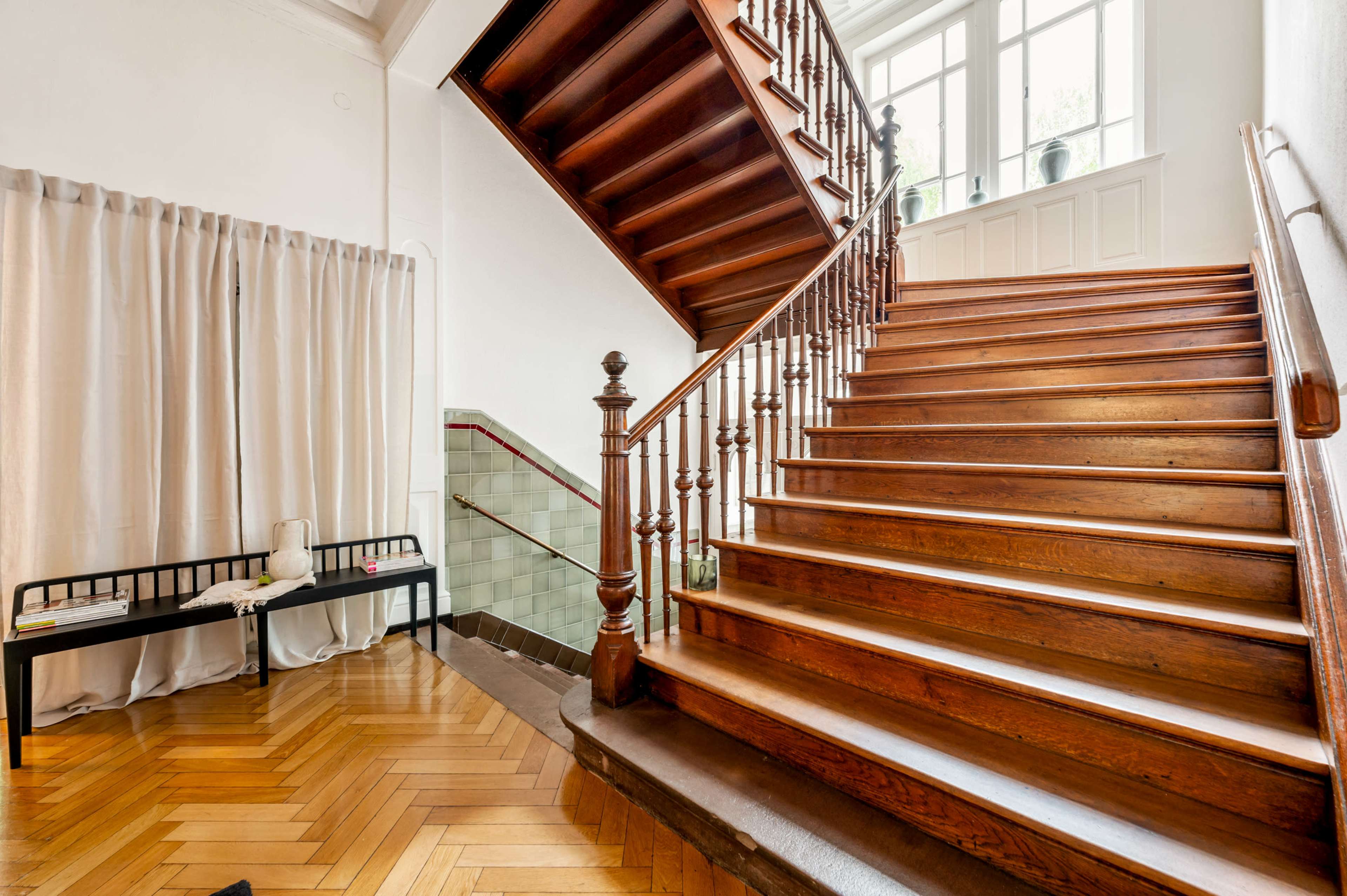 The image depicts a wooden staircase with a bannister, leading to an upper level, alongside a bench and a large window with natural light.