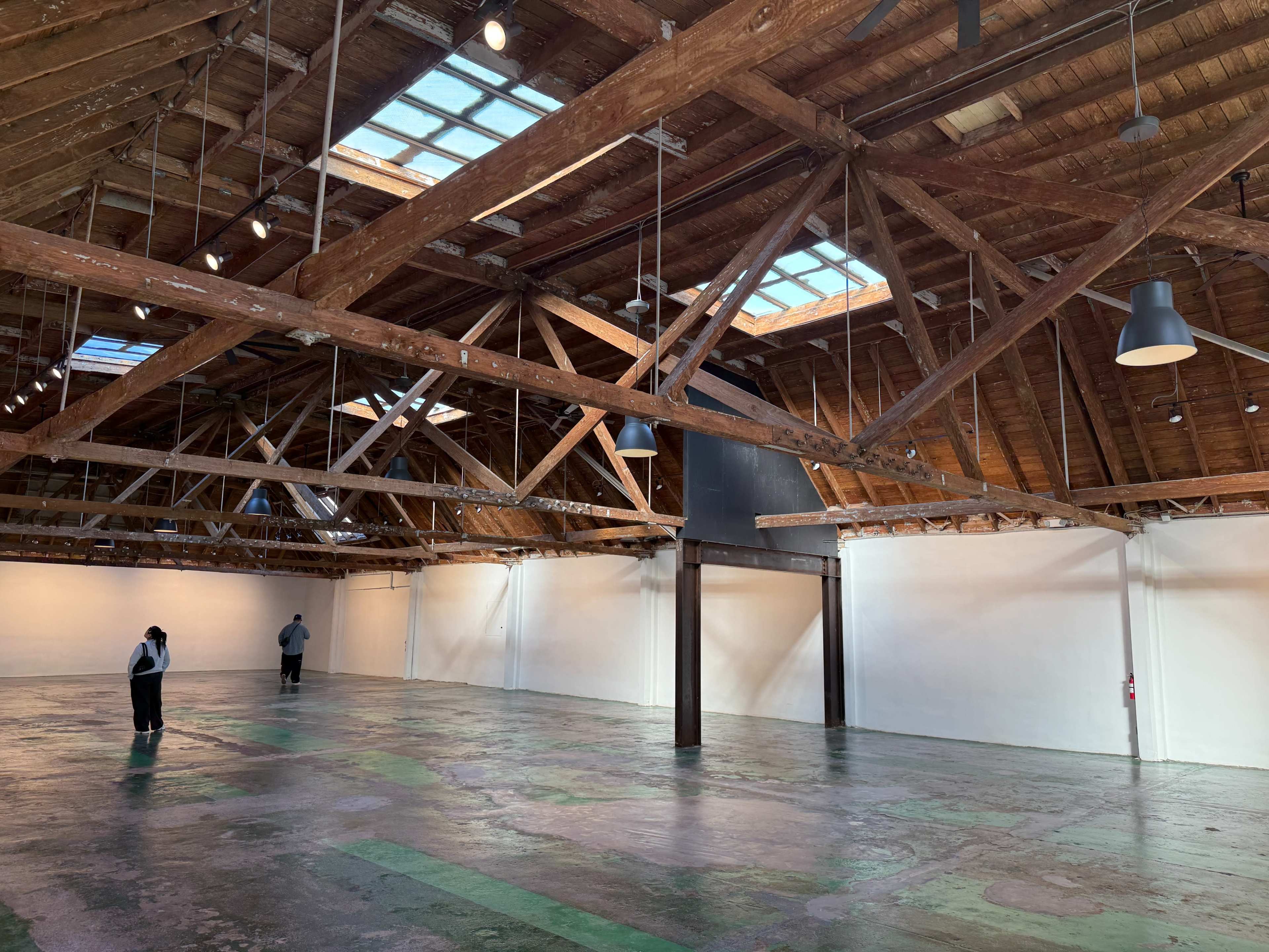 The image shows a spacious, empty warehouse with exposed wooden beams and skylights, featuring a concrete floor and two people standing at opposite ends of the room.