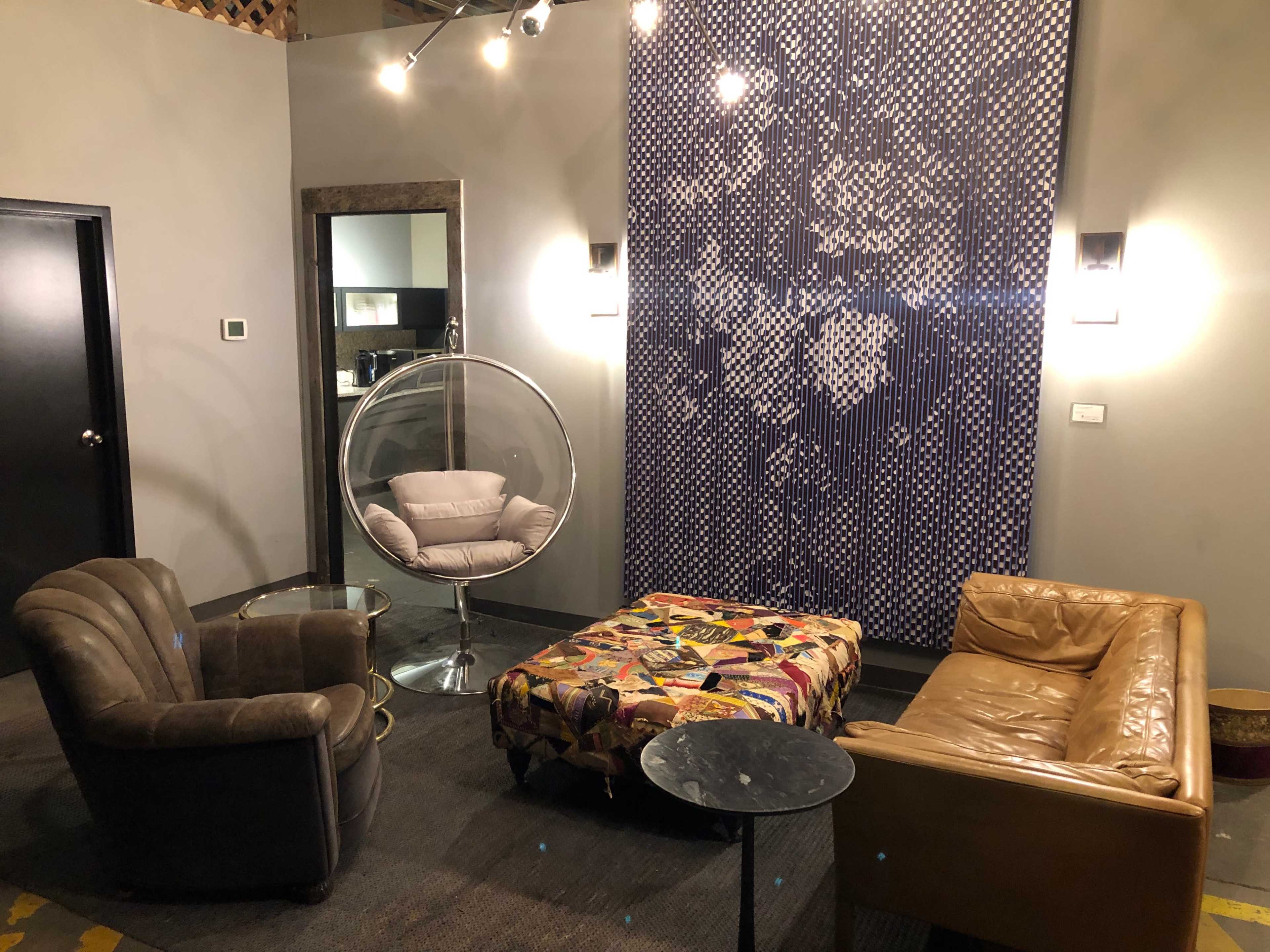A modern lounge area featuring two armchairs, a hanging bubble chair, a colorful ottoman, and a patterned wall decoration.