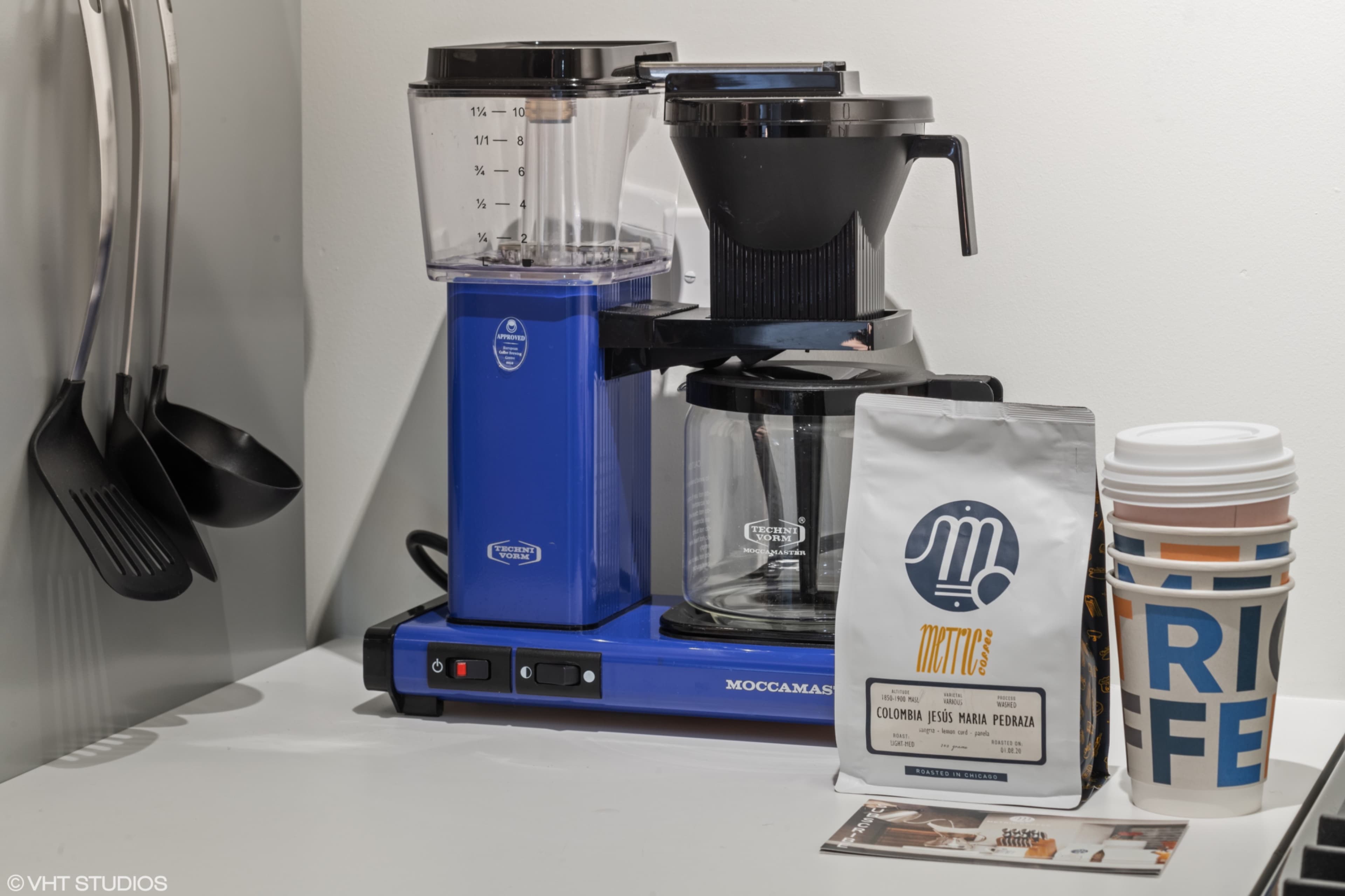 A blue coffee maker sits on a countertop next to a bag of coffee and several disposable cups.