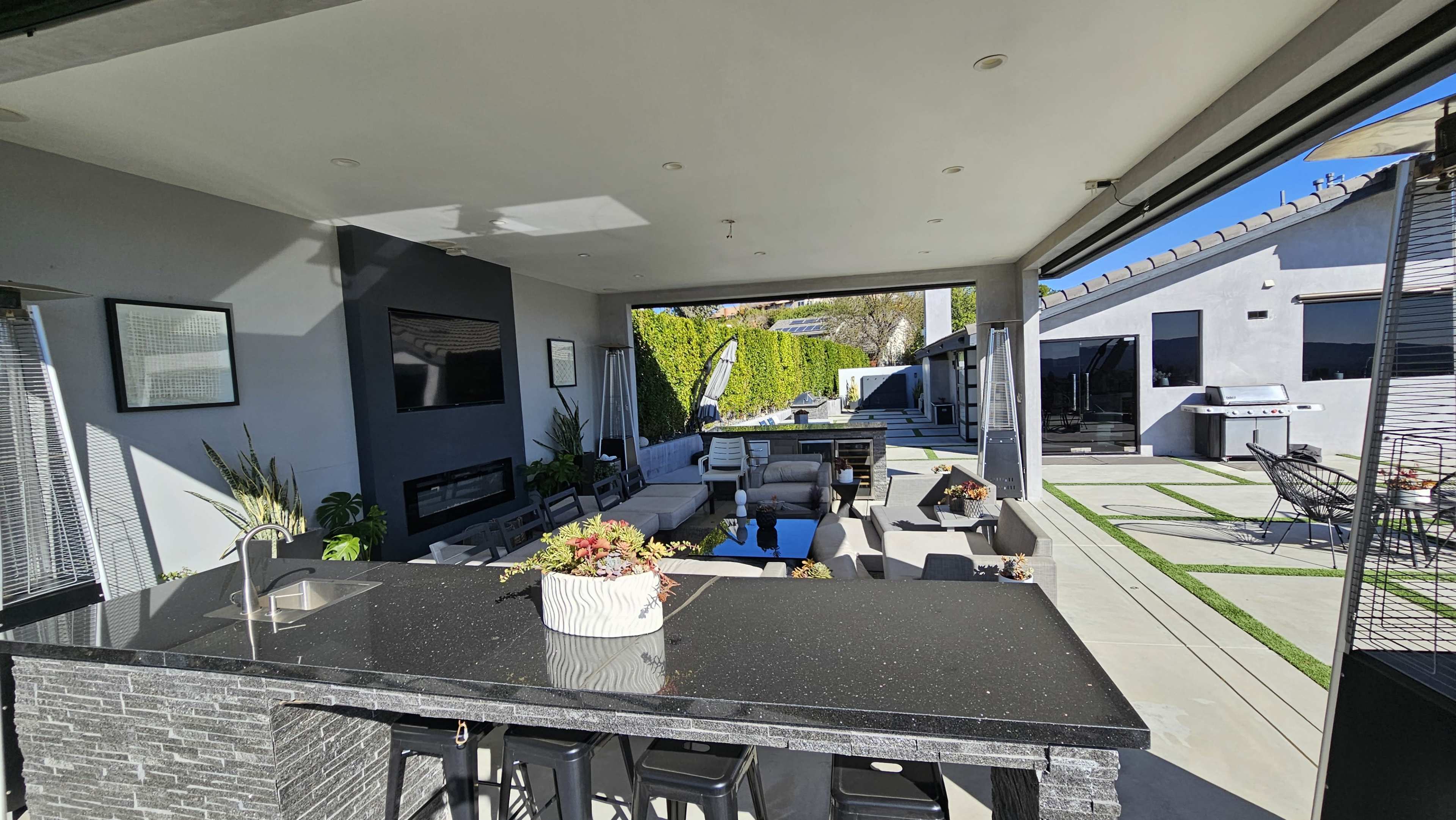 The image shows a modern outdoor living space featuring a kitchen island, seating area, and a backyard with landscaping and a barbecue area.