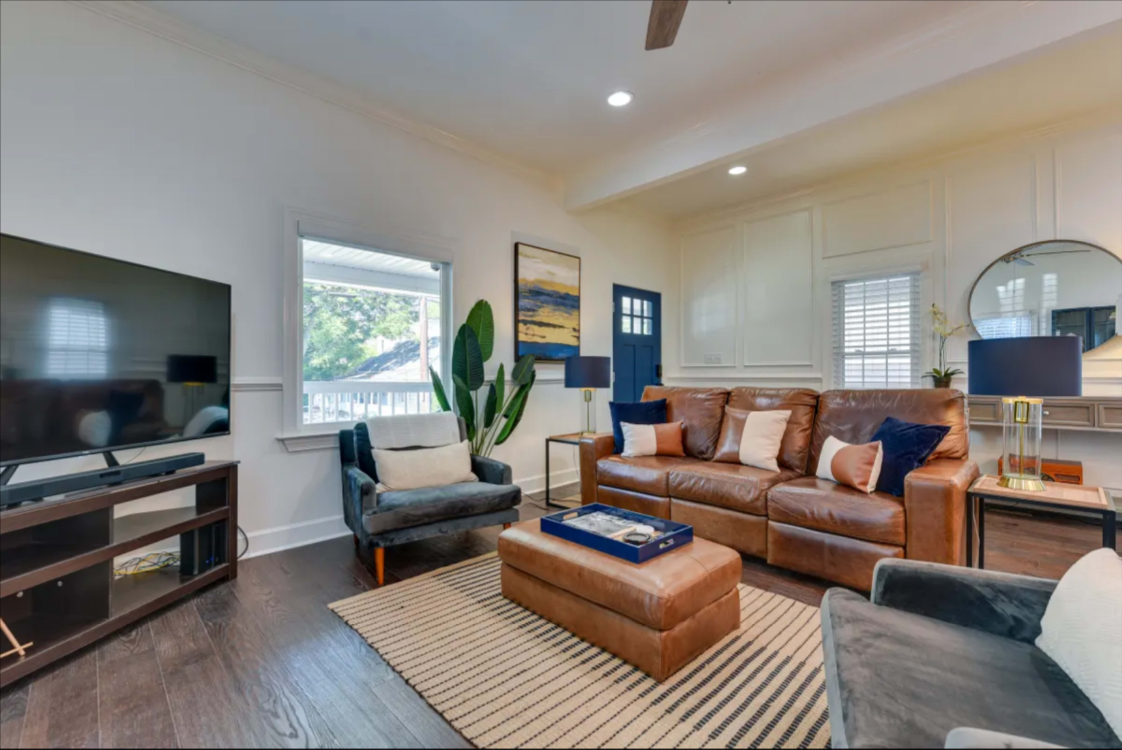 A modern living room features a brown leather sofa, two chairs, a television console, and large windows with natural light.