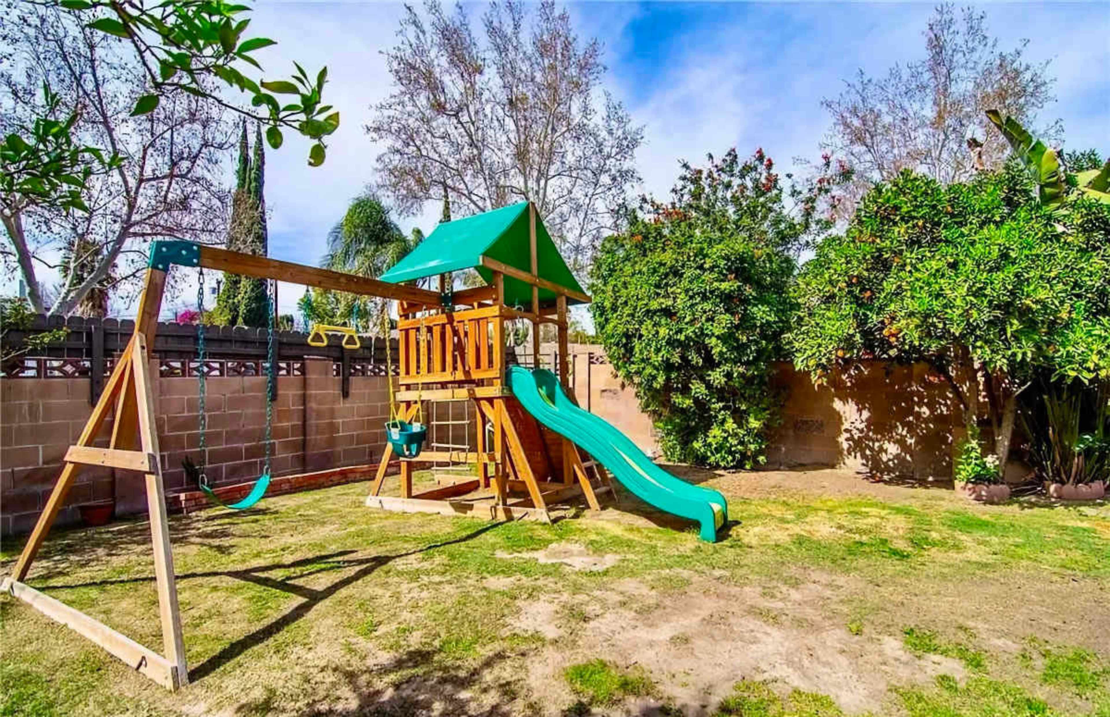 A wooden playground set with a slide, swings, and a fort is installed in a grassy backyard surrounded by trees and shrubs.