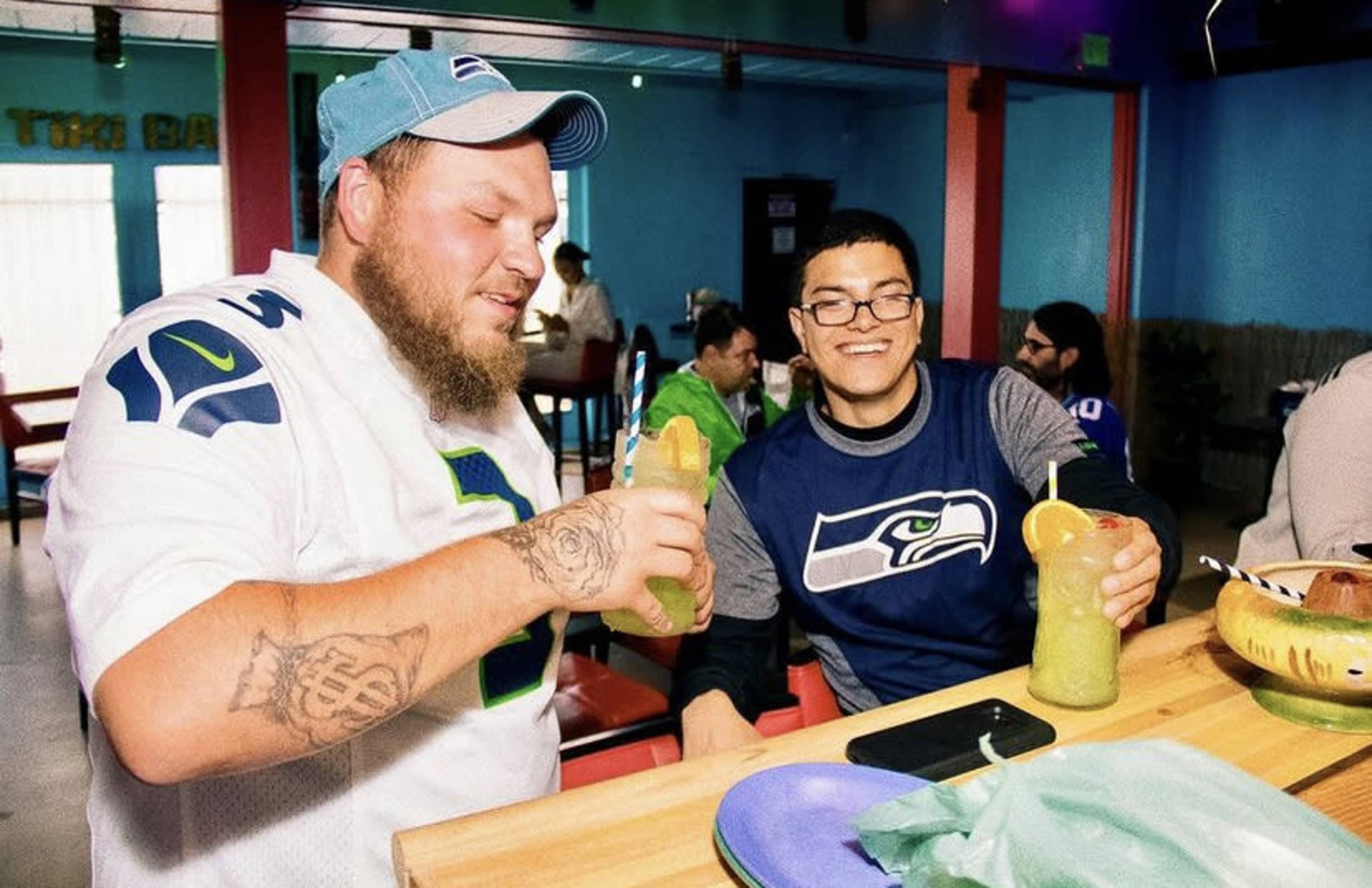Two men sit at a brightly colored table in a restaurant, holding large green drinks and wearing Seattle Seahawks apparel.