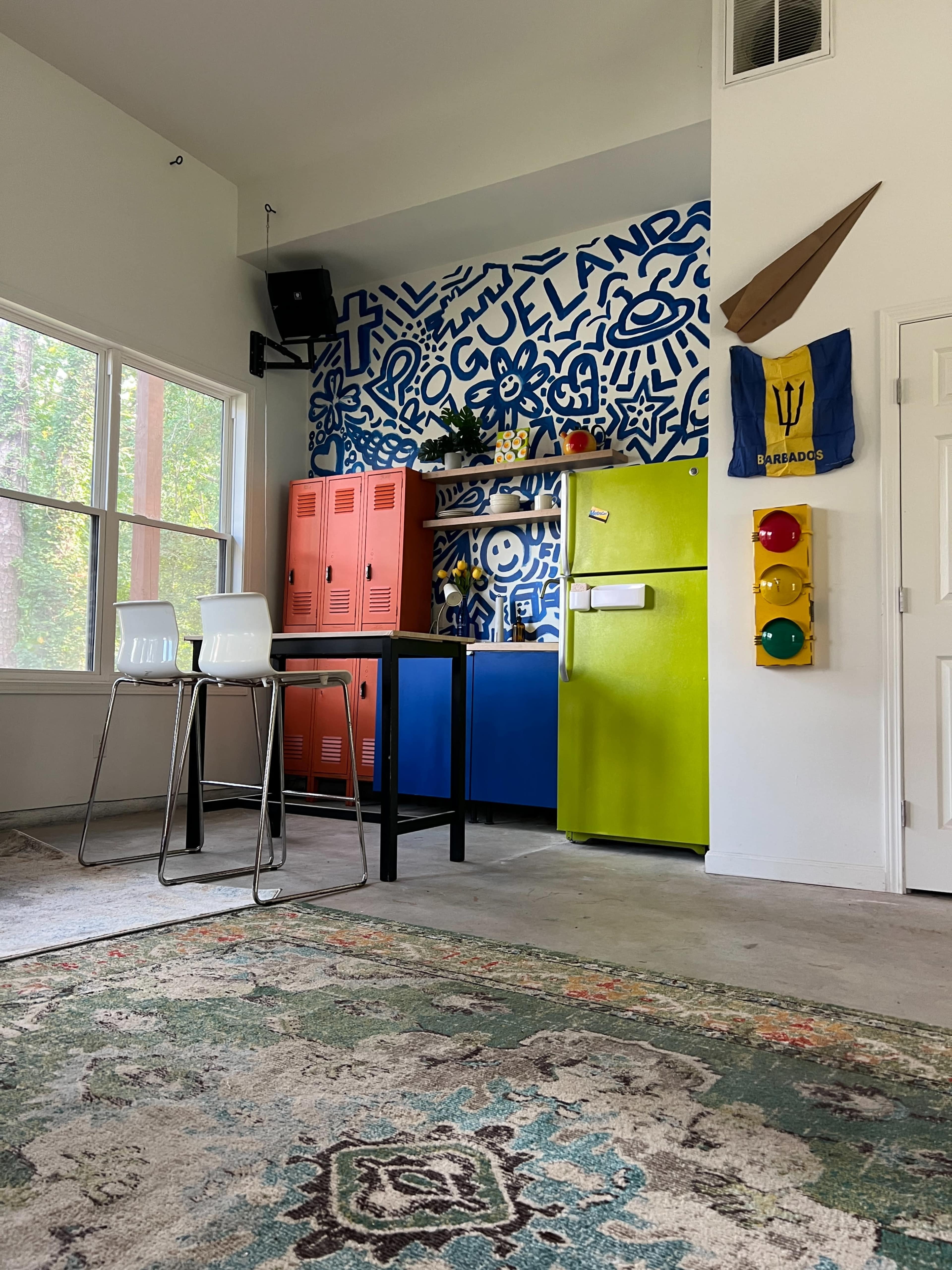 The image shows a brightly colored kitchen area featuring blue and green appliances, orange lockers, and a patterned rug on the floor.