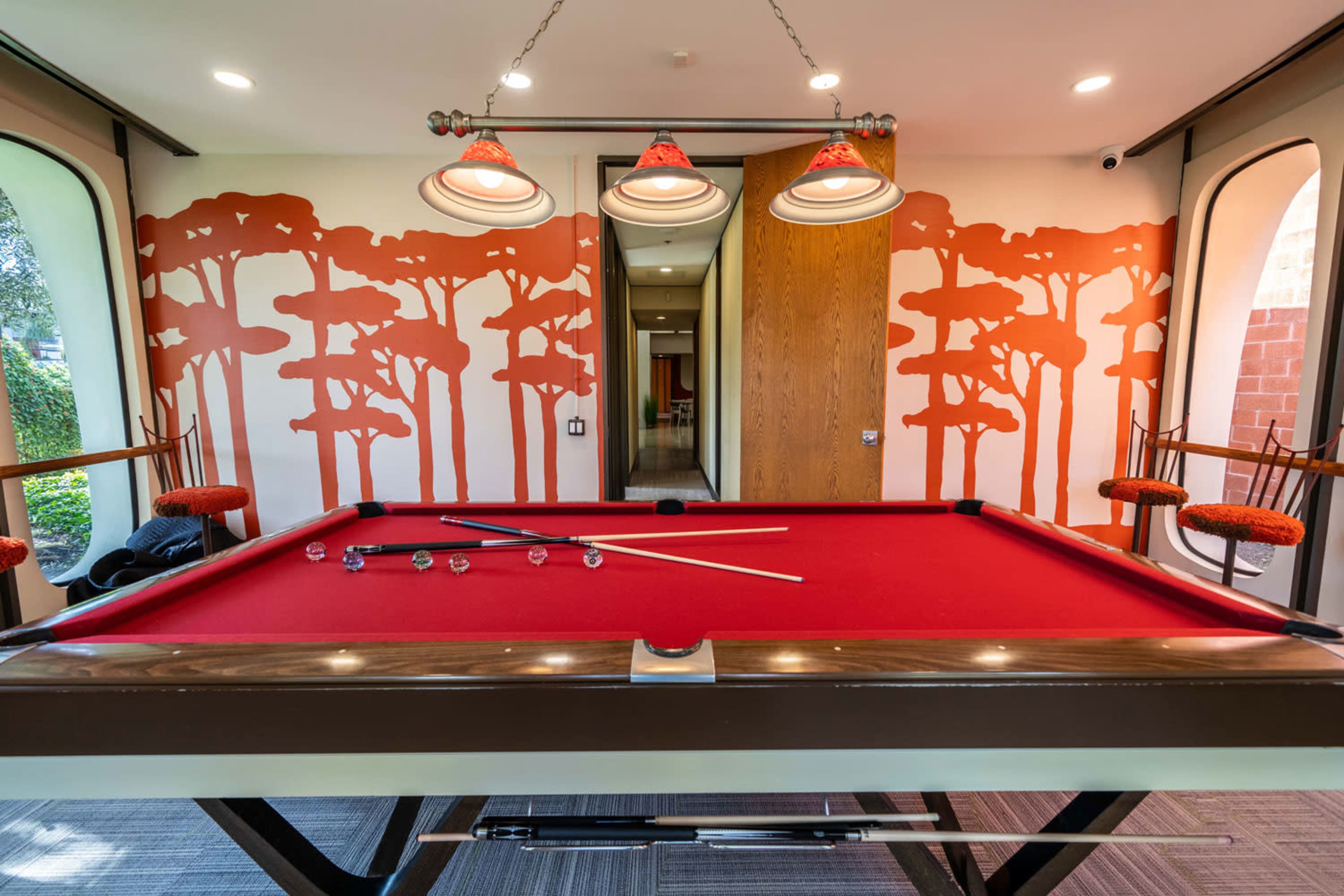 A red pool table, with cues and balls, is set up in a room featuring orange tree-patterned wallpaper and modern lighting fixtures.