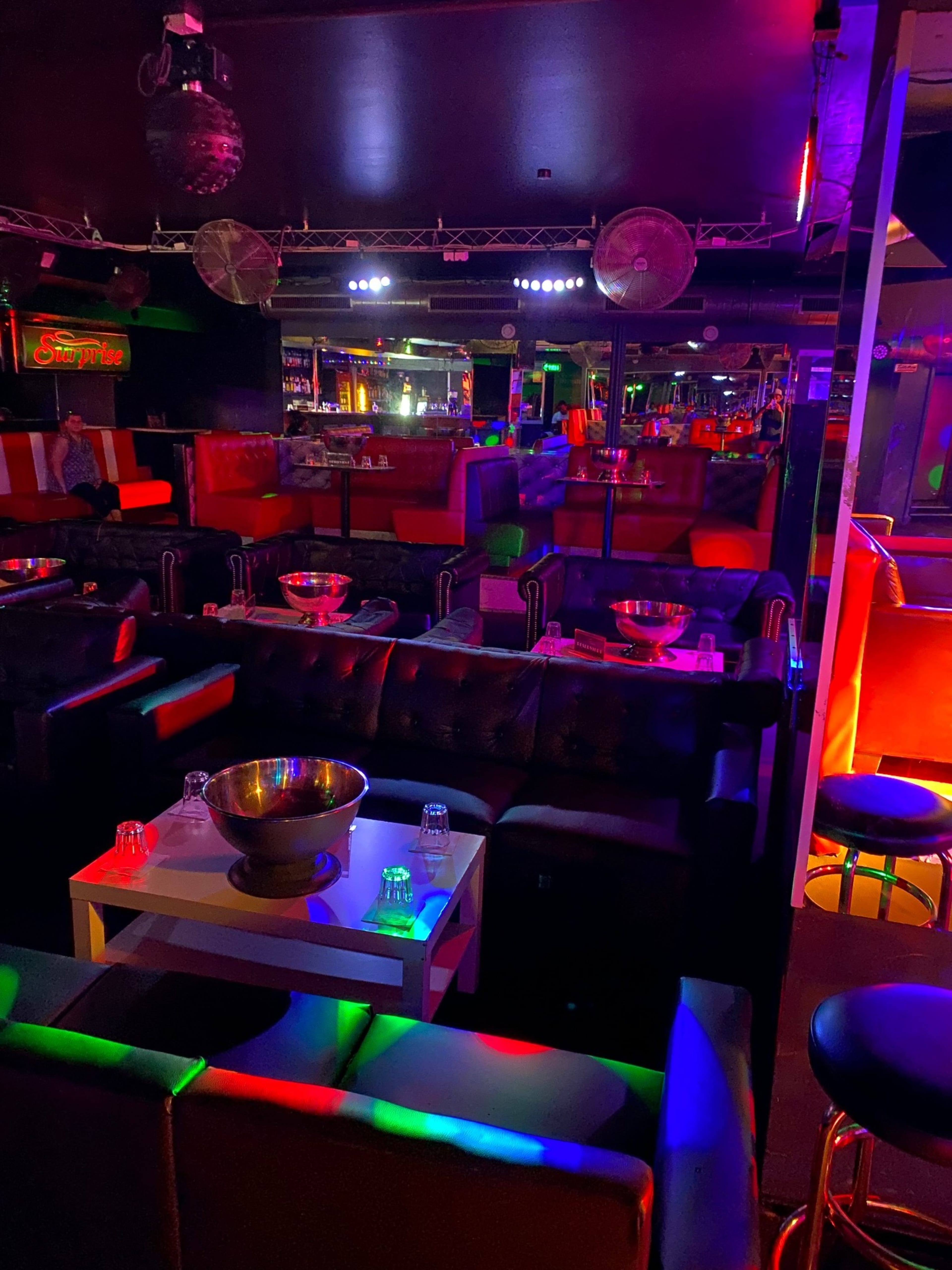 The image shows the interior of a dimly lit lounge or nightclub featuring black leather seating, colorful lighting, and several tables set with bottles and glasses.
