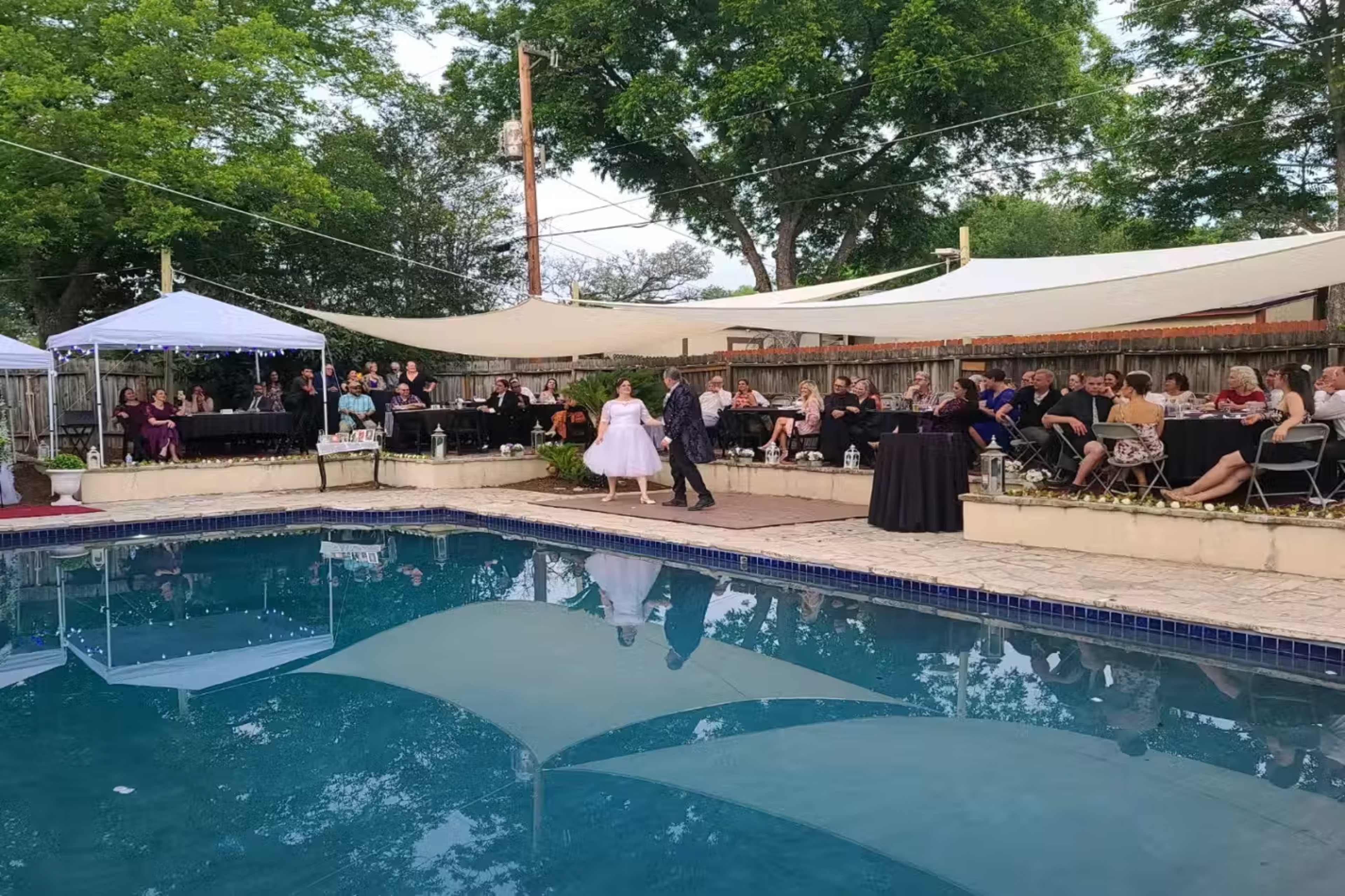 A couple dances by the poolside at a wedding reception, while guests watch from seated tables under shaded canopies.