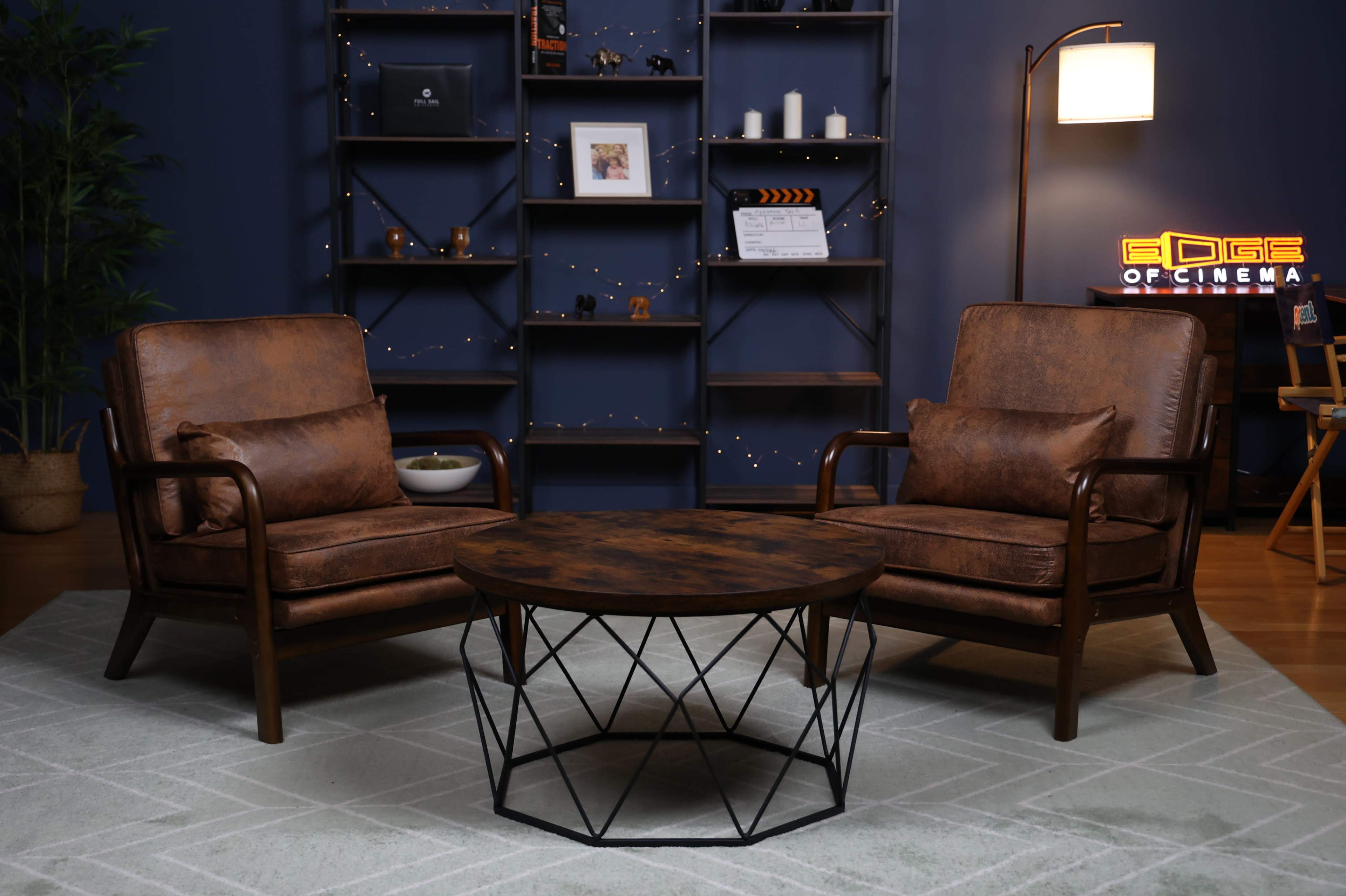 The image shows a cozy seating area with two brown armchairs facing a round coffee table, set against a blue wall with decorative shelving and soft lighting.
