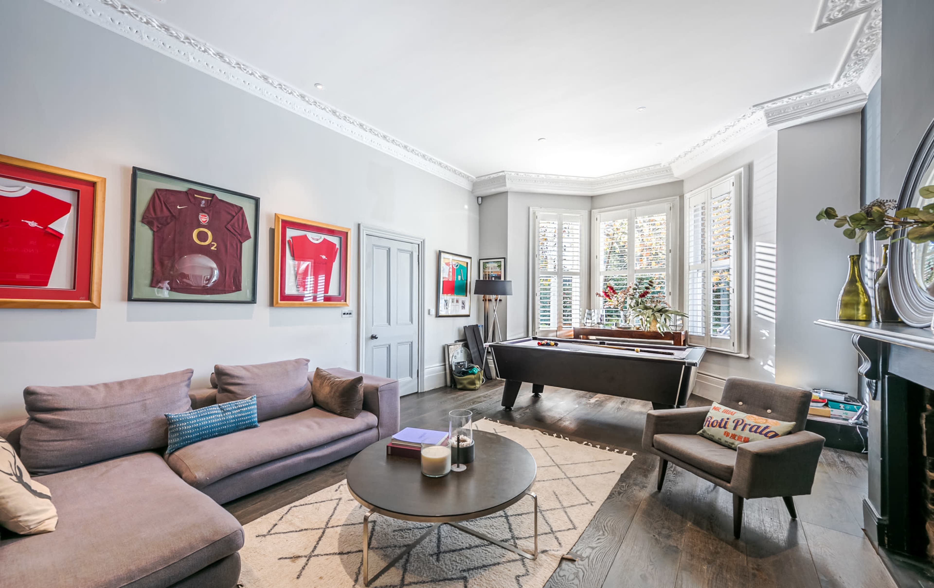 A living room features a gray color scheme with framed jerseys on the walls, a sectional sofa, and a window with decorative shutters.