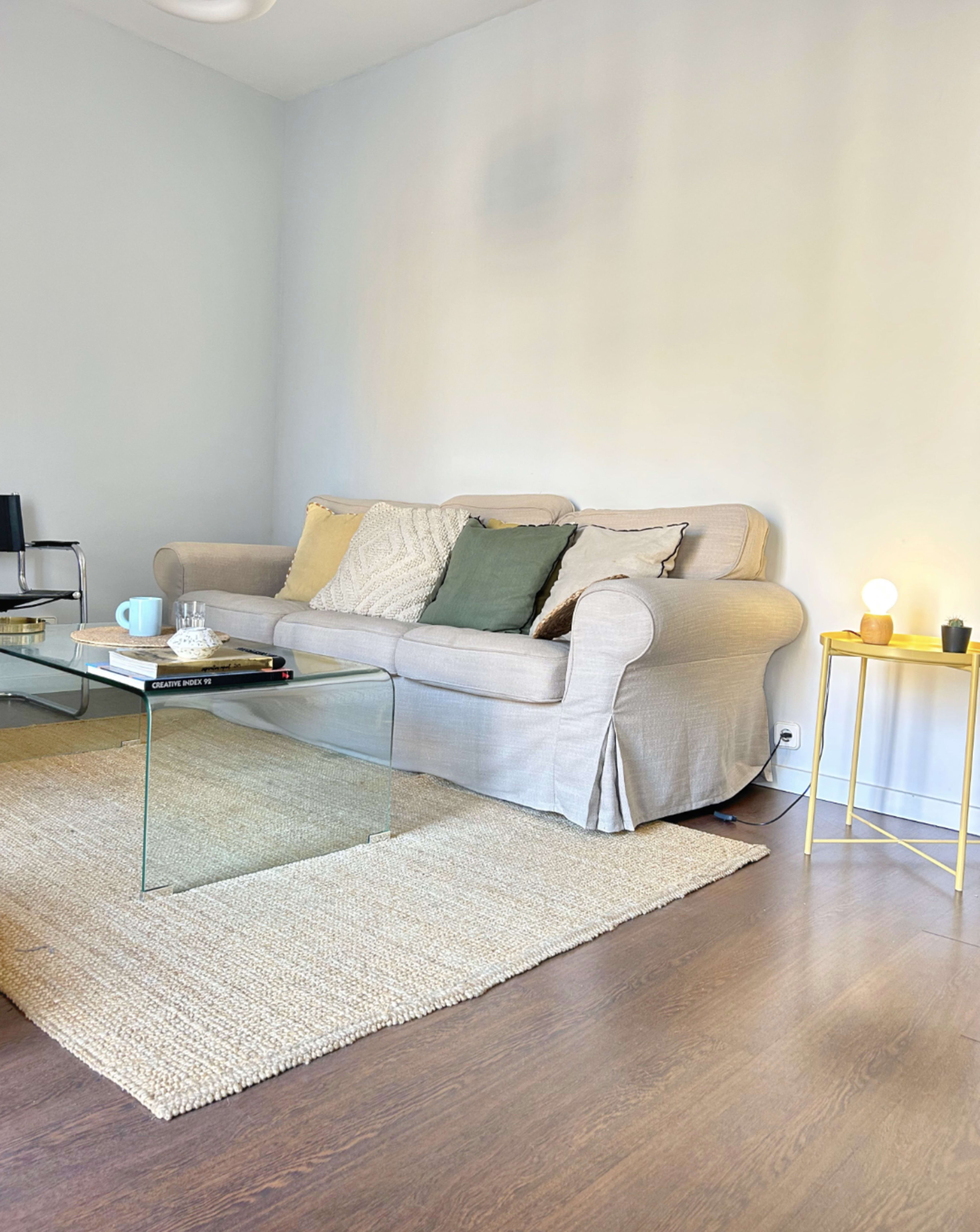 A light-colored sofa with decorative pillows sits in a minimalistic living room beside a glass coffee table and a textured area rug.