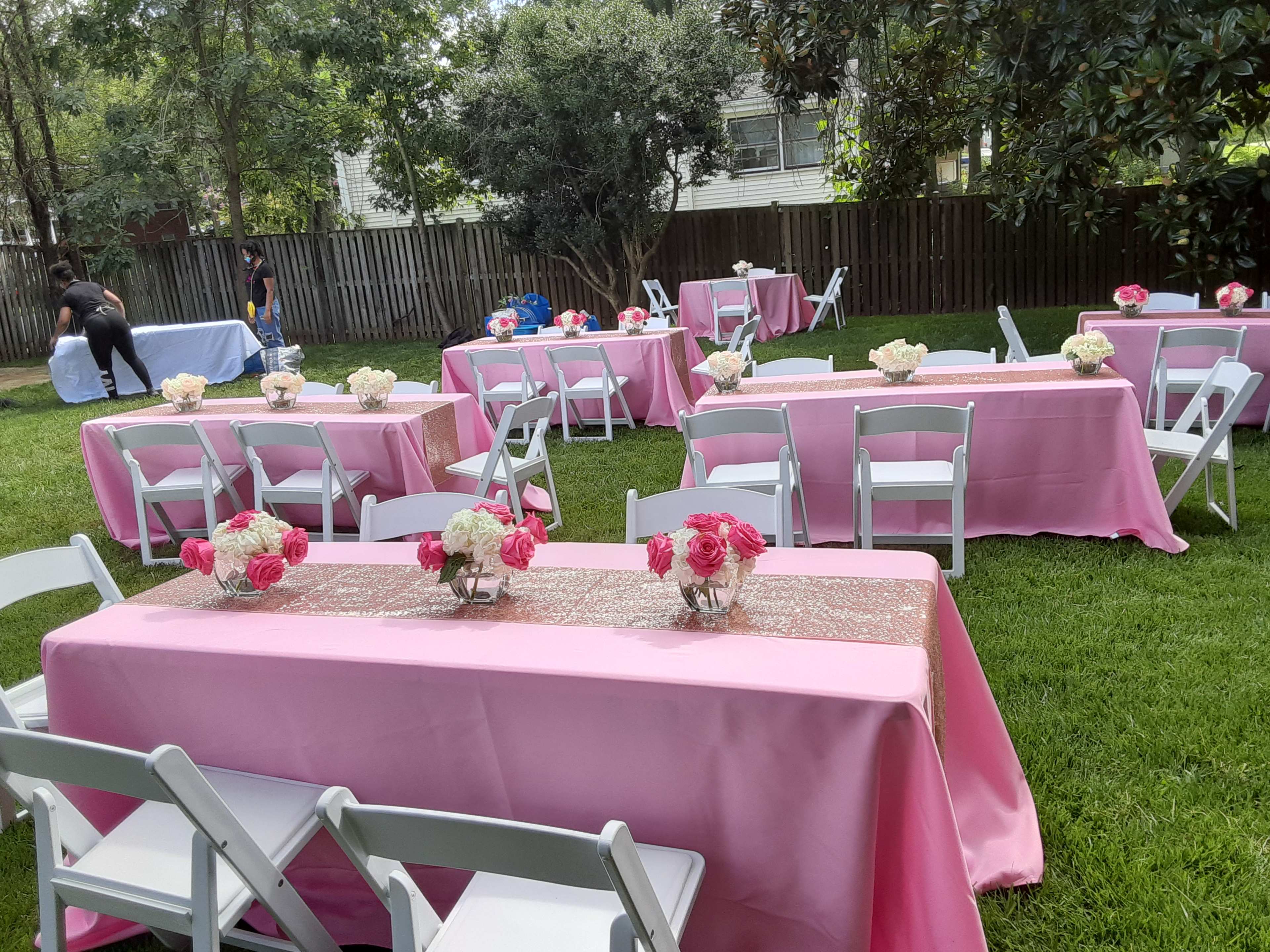 A garden setup with several tables covered in pink tablecloths and adorned with flower arrangements, surrounded by greenery.