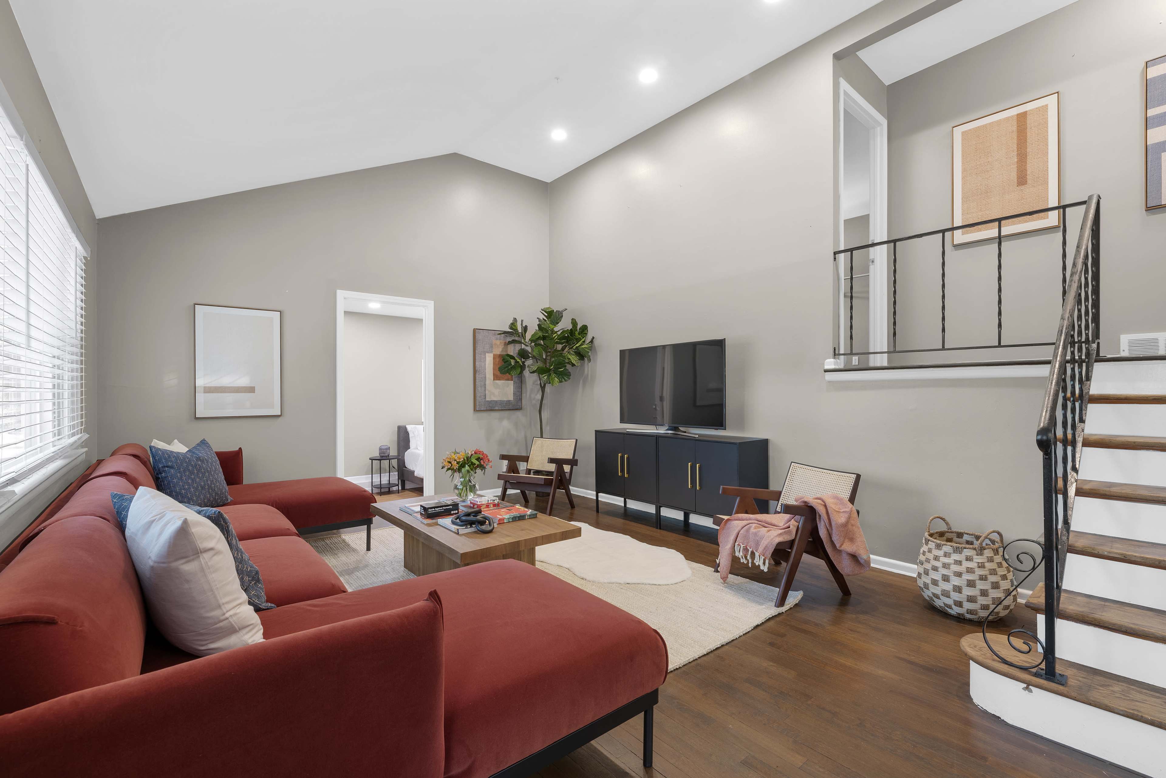 The bright living room features a red sectional sofa, a wooden coffee table, a television stand, and a staircase leading to an upper level.