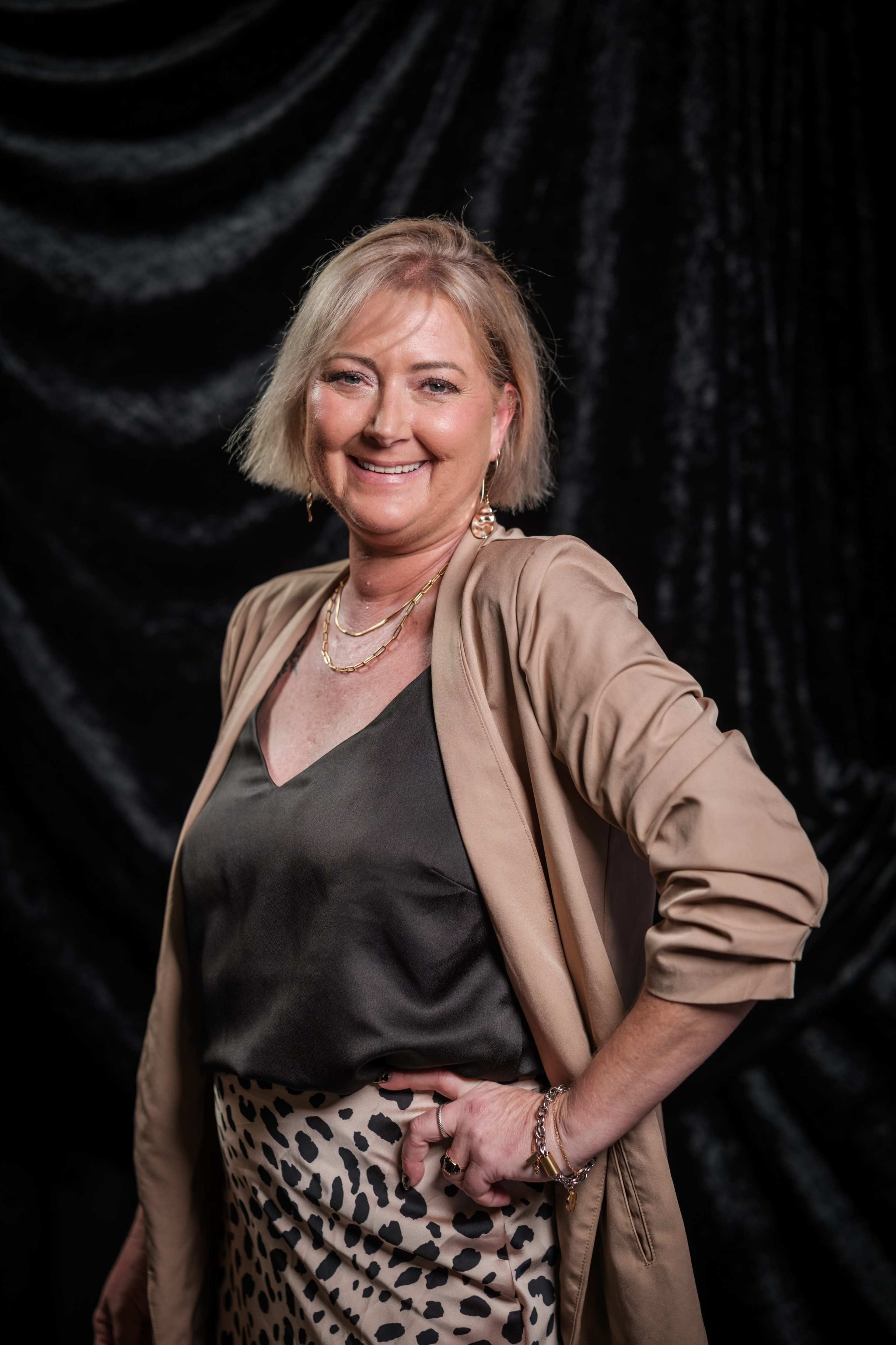 A woman with short blonde hair poses confidently in front of a dark, textured backdrop while wearing a stylish outfit.