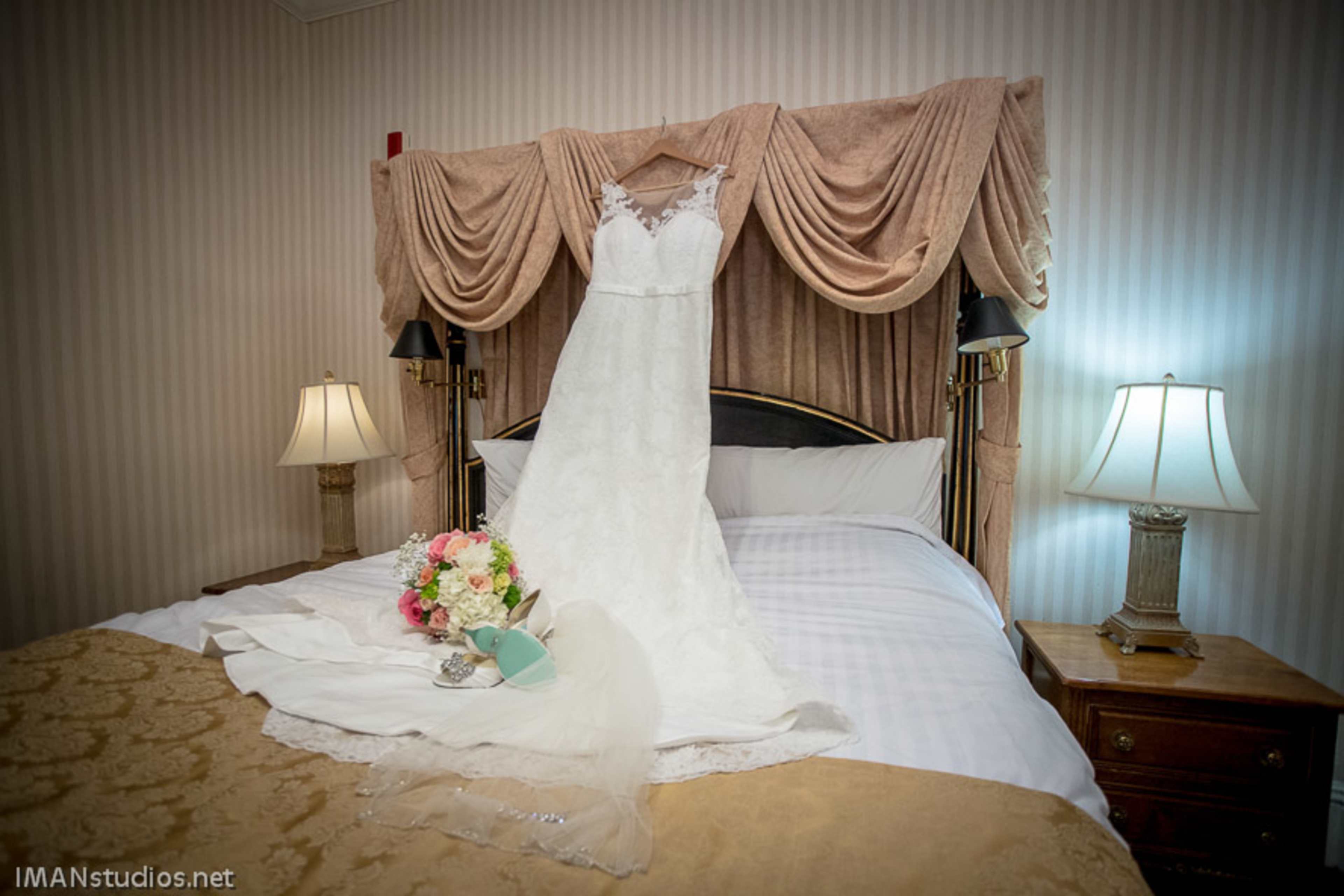 A wedding dress is displayed on a bed with a bouquet and accessories arranged nearby.
