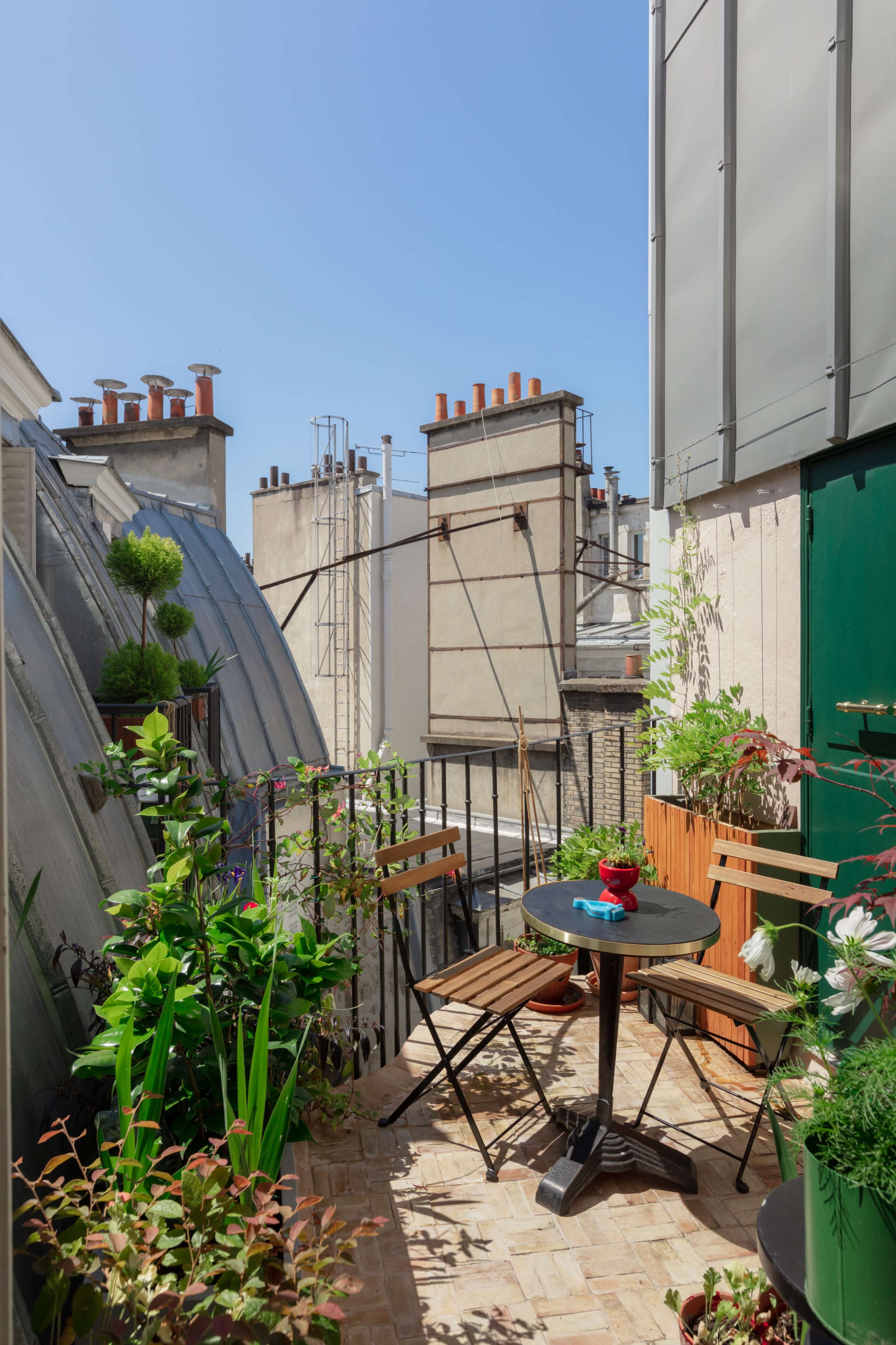A small balcony terrace features potted plants, a round table, and two chairs, set against a backdrop of buildings with chimneys and clear blue sky.