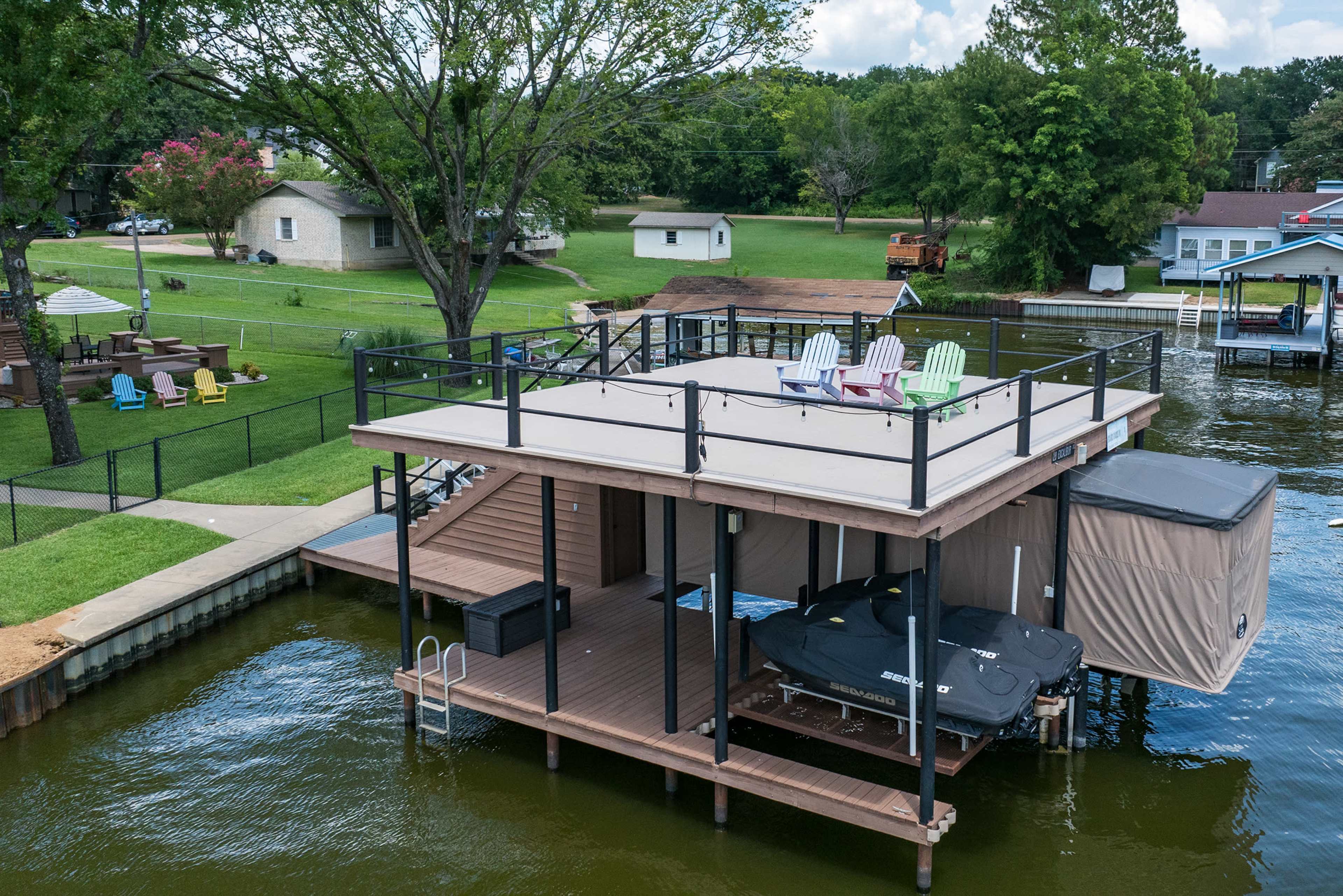 A wooden boat dock features a shaded seating area with colorful chairs and a boat lift on a calm lake surrounded by green trees and houses.