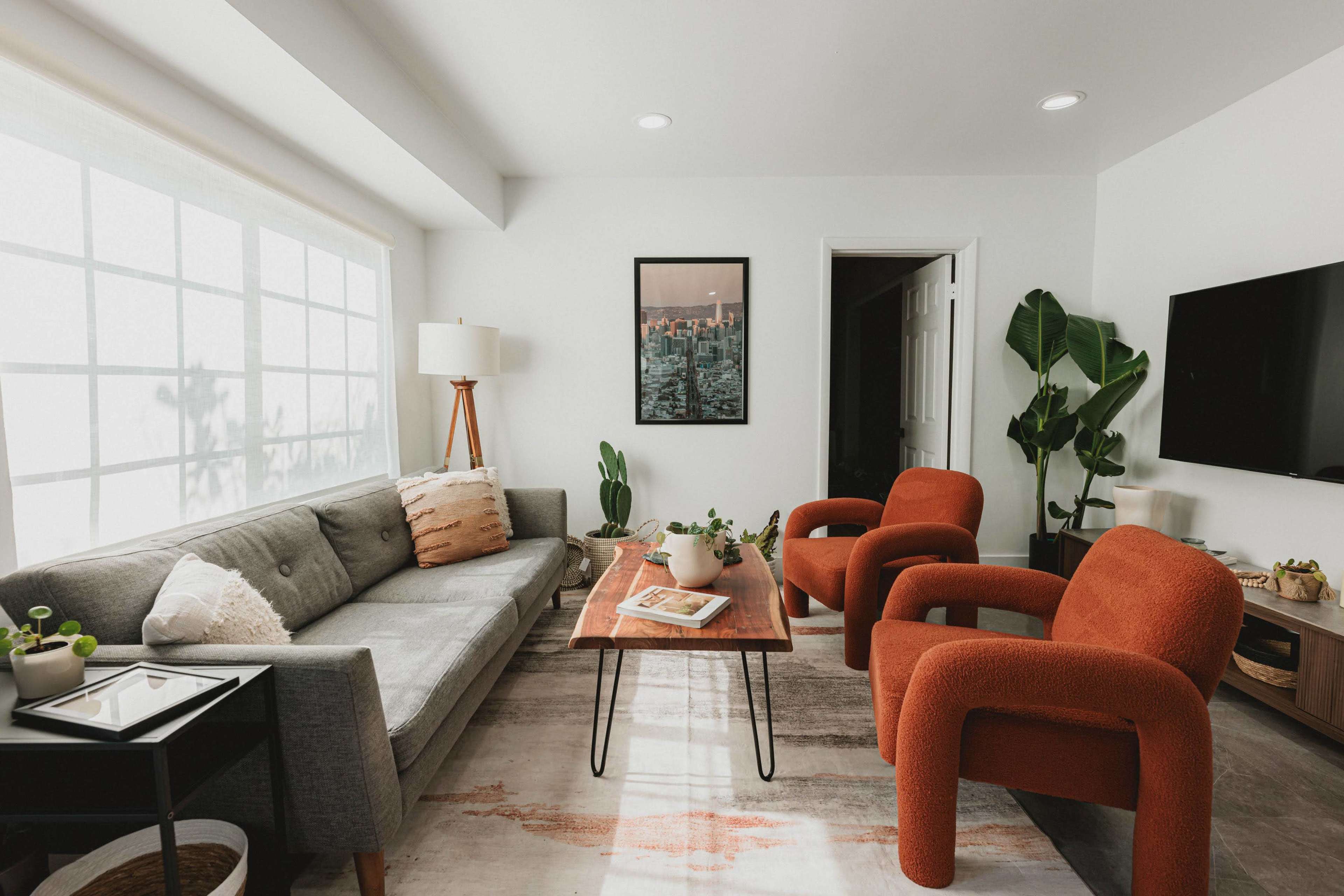 A modern living room features a gray sofa, two orange chairs, a wooden coffee table, and large windows allowing natural light to brighten the space.